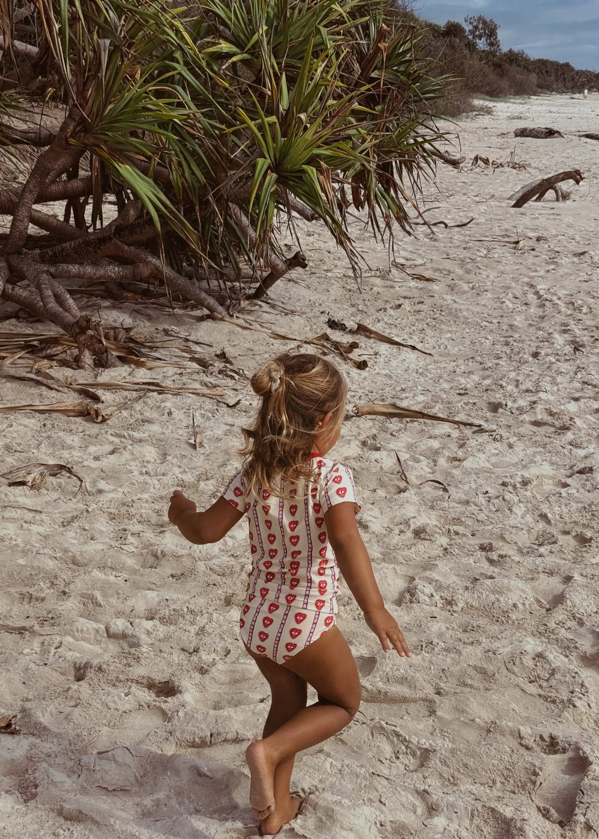 A young girl with blonde hair in a ponytail walks barefoot on a sandy beach, viewed from behind. She is wearing a white short-sleeve one-piece swimsuit with a red heart pattern.
