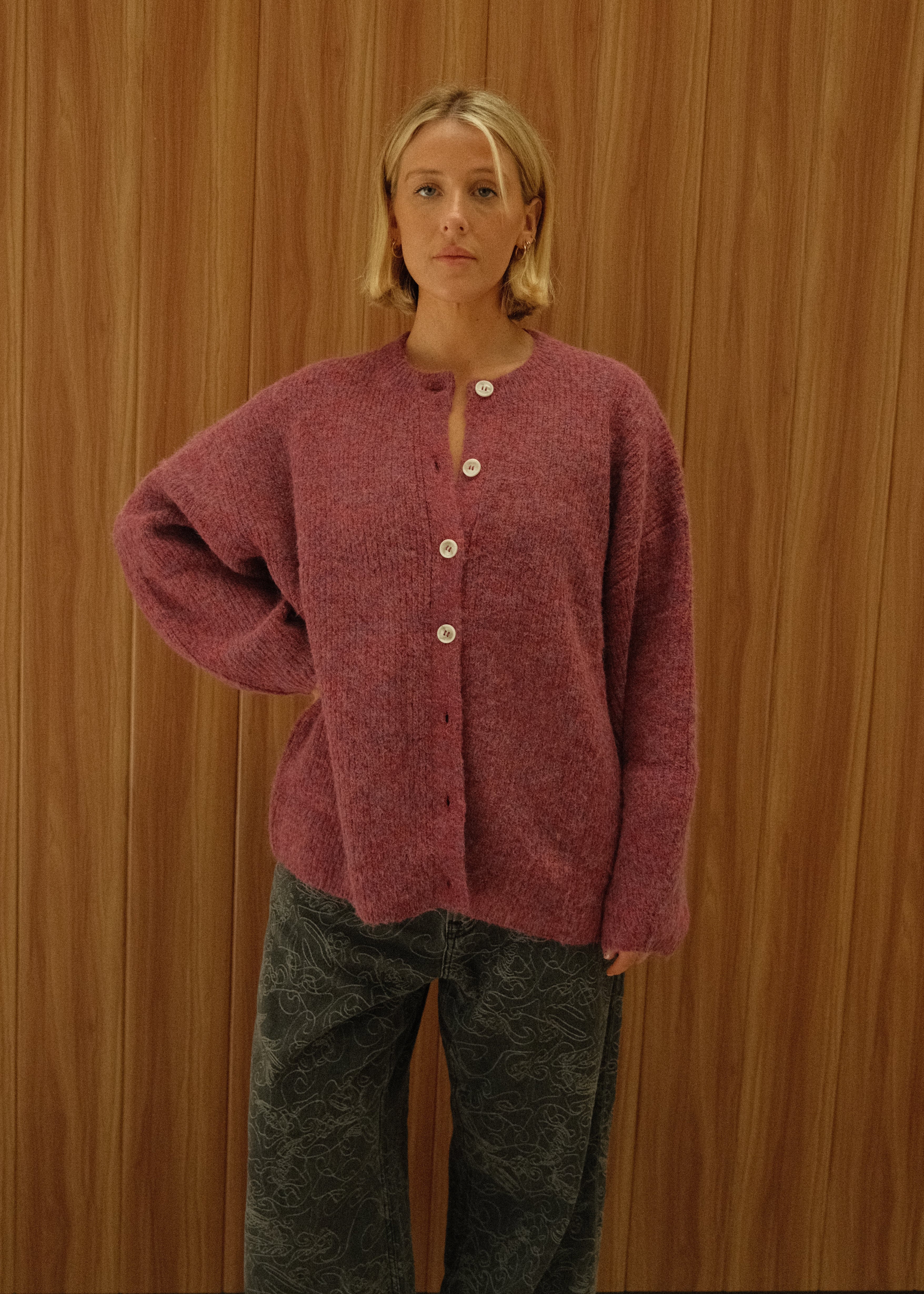 A woman wearing a dusty rose pink button-up cardigan and dark gray patterned pants, standing against a wooden wall background.