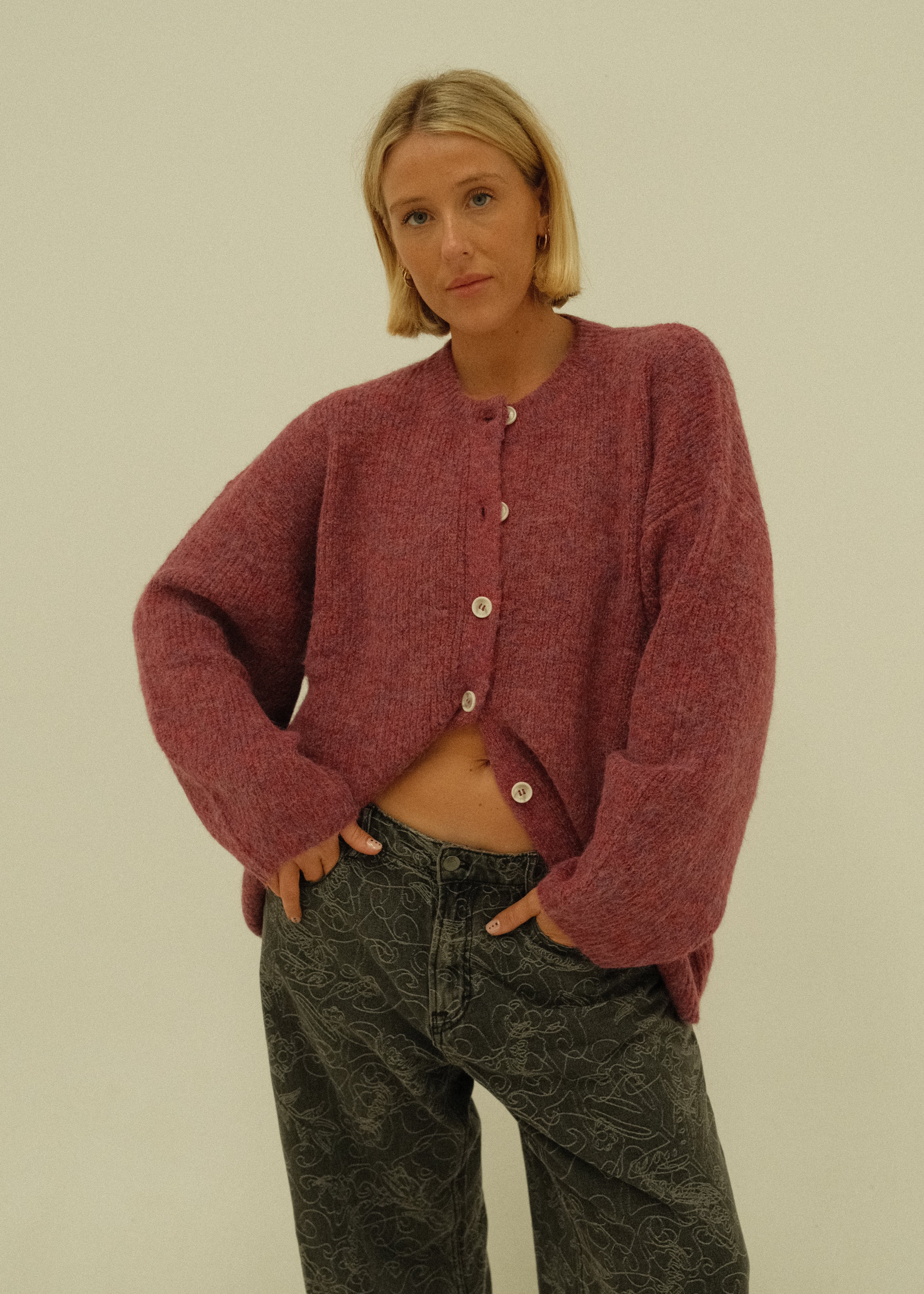 A woman wearing a textured, heathered dusty rose cardigan and dark gray patterned jeans, photographed against a plain off-white background.