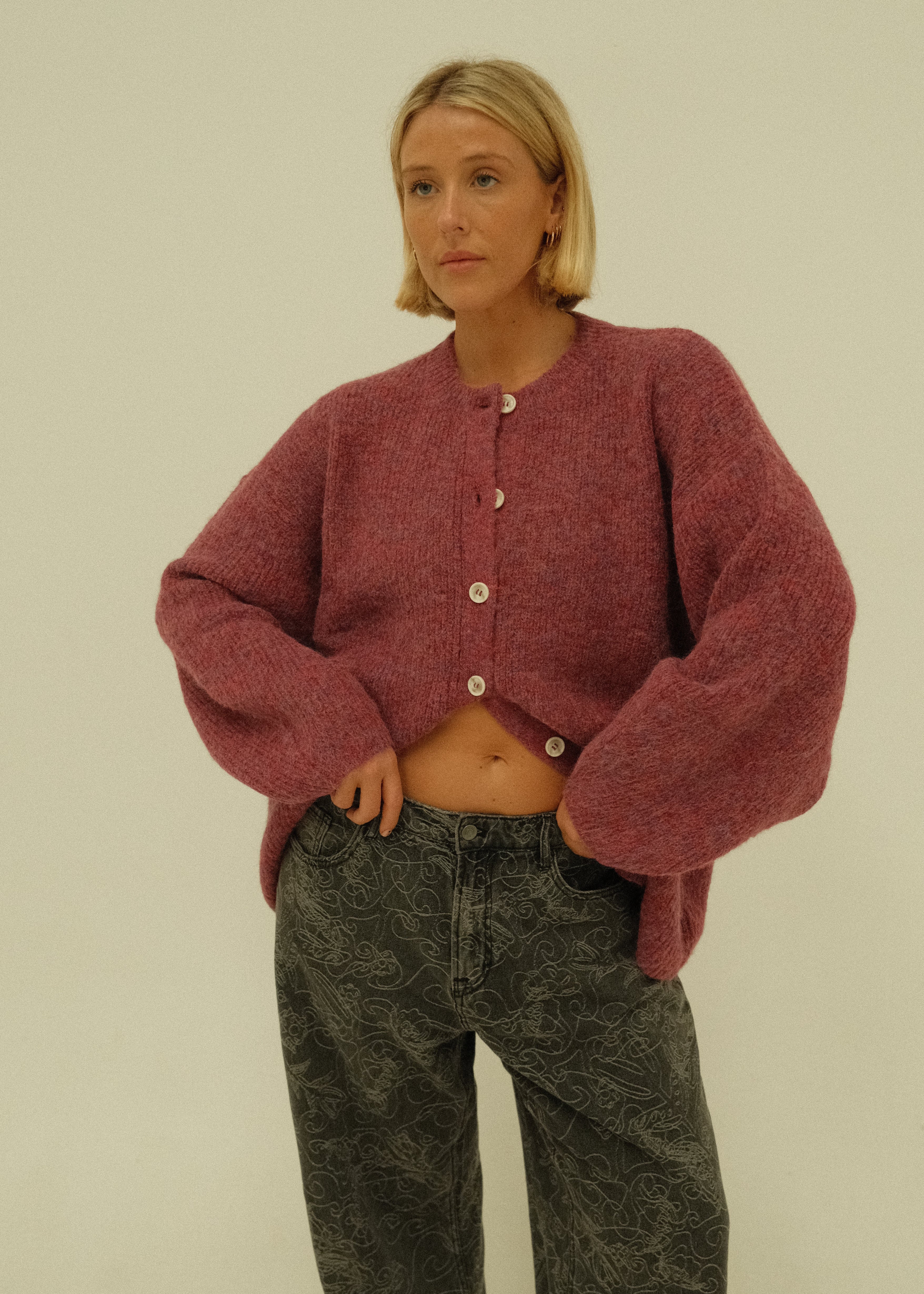 A person wearing a fuzzy dusty rose cardigan and dark gray jeans, posed against a plain off-white background.