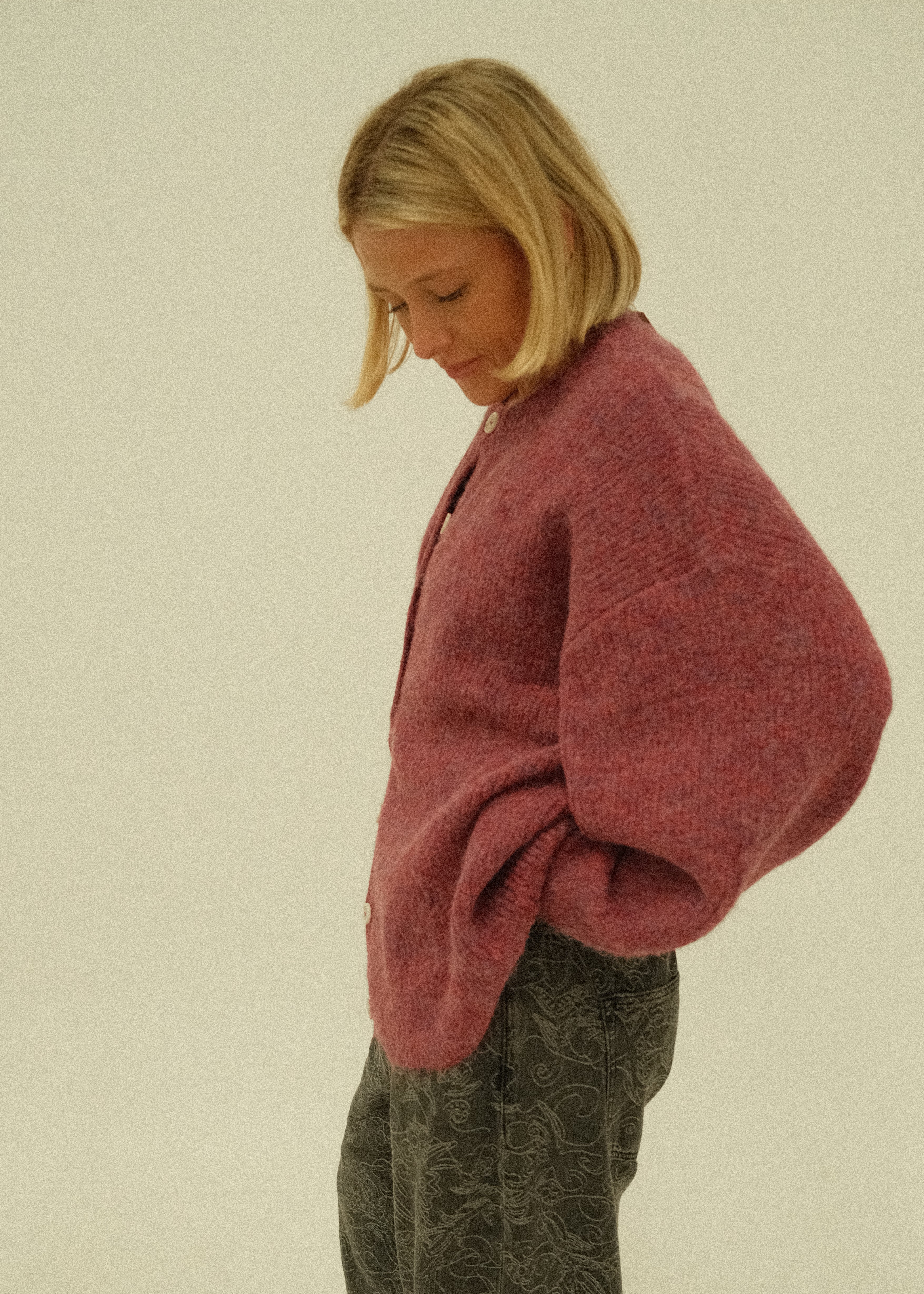 A woman lying down wearing a dusty rose pink knit sweater and dark gray textured pants, photographed against a plain off-white background.