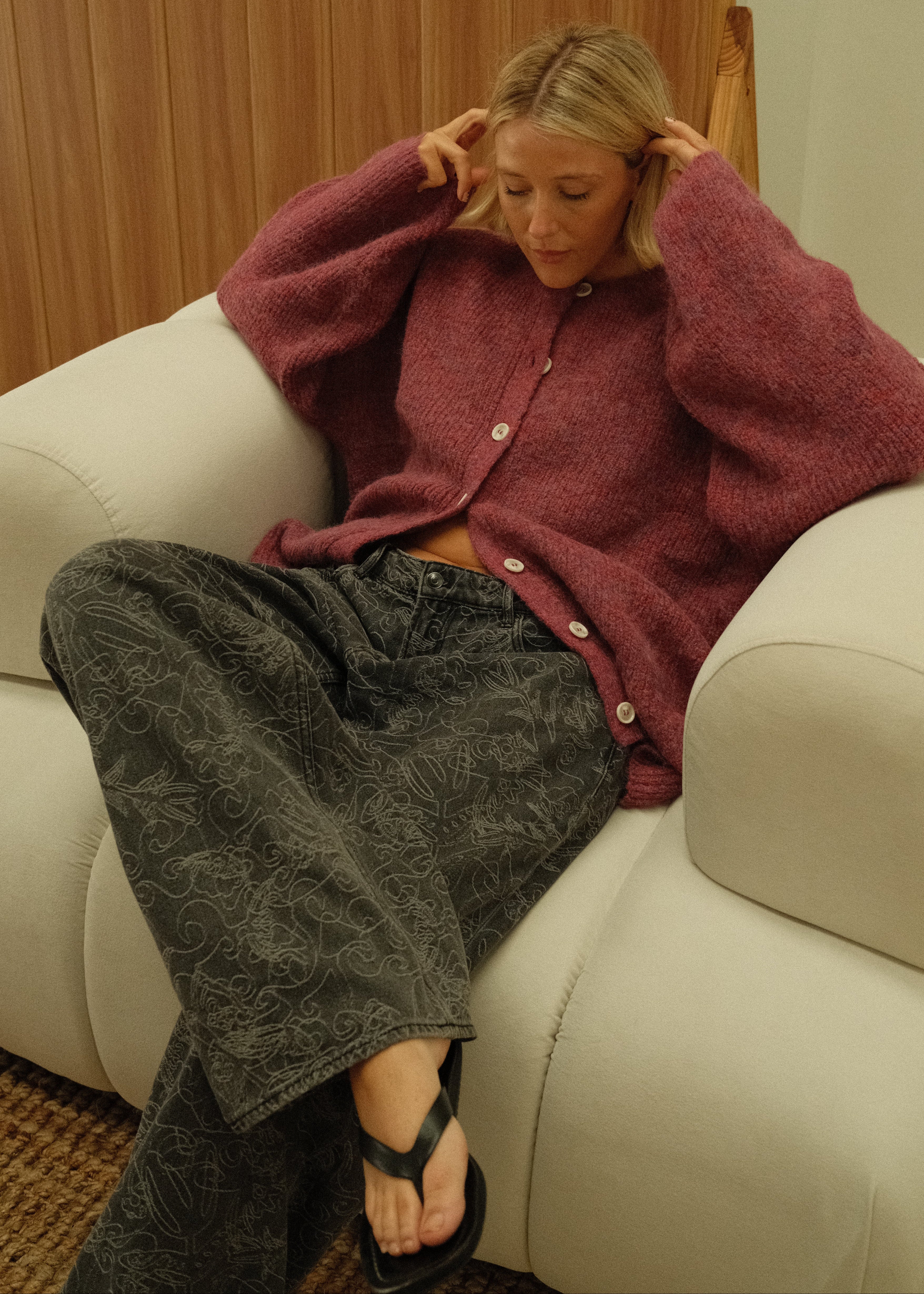 A woman reclining on a cream-colored sofa in a home setting, wearing a dusty rose cardigan and dark gray patterned pants.