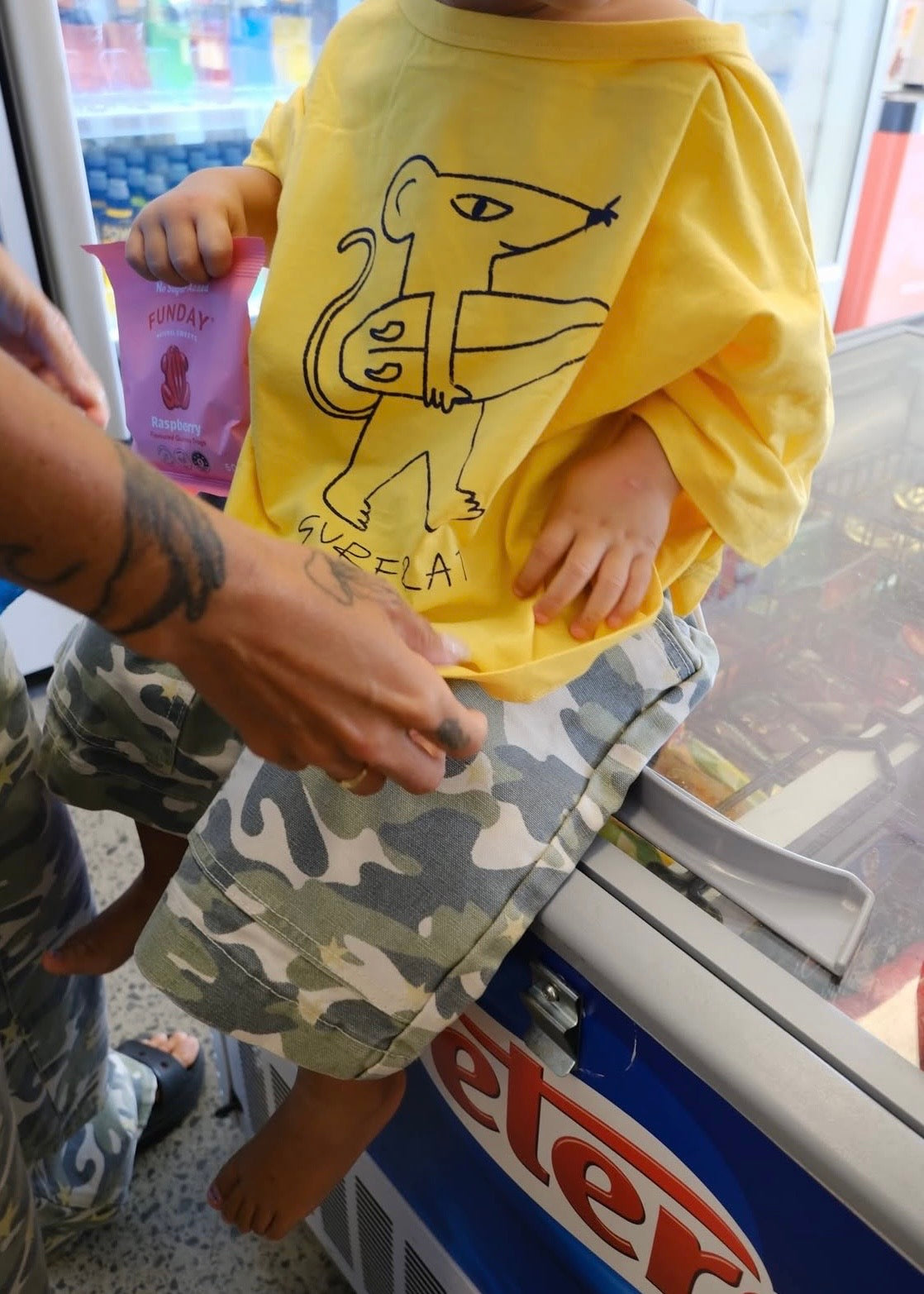A child wearing a bright yellow t-shirt with a black line art graphic of a mouse and the word 'GUEILA' paired with camouflage shorts, sitting on a freezer display in a store while an adult's arm assists.