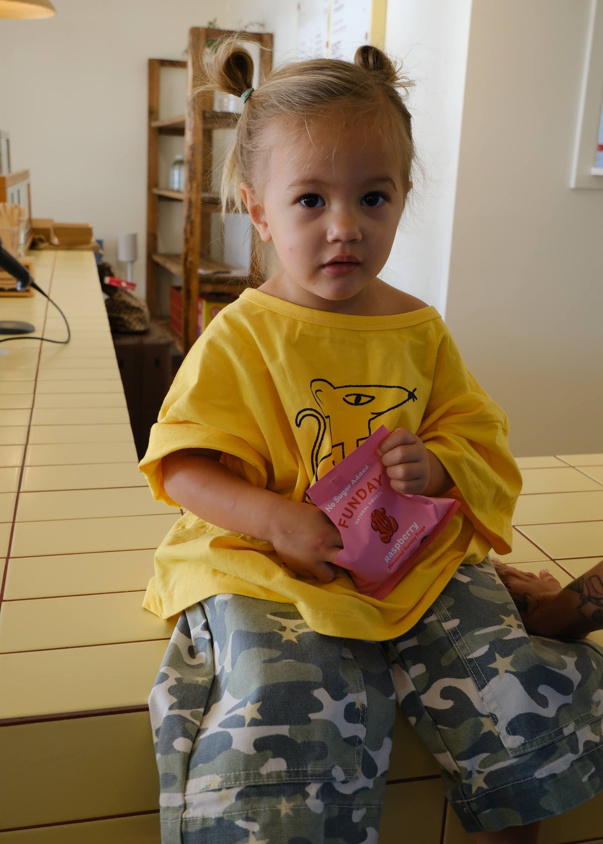A young child with blonde pigtails sits on a tiled counter wearing a yellow t-shirt with a cartoon graphic and blue-gray camouflage shorts. The child is holding a pink bag of Famosa snacks in a kitchen-like setting.