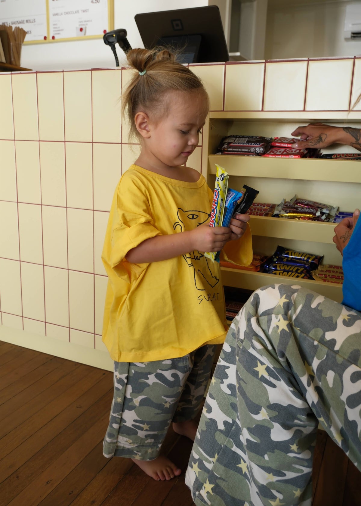 A young girl with her hair in a ponytail stands in a shop or cafe setting, wearing a yellow t-shirt and camouflage pants while holding colorful candy bars. A counter with a laptop and shelves of snacks is visible in the background.