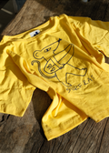 A bright yellow short-sleeve t-shirt with a black line art graphic of a surfboard cat and the text 'SURF & 1' printed on the front, laid flat on a rustic wooden surface.