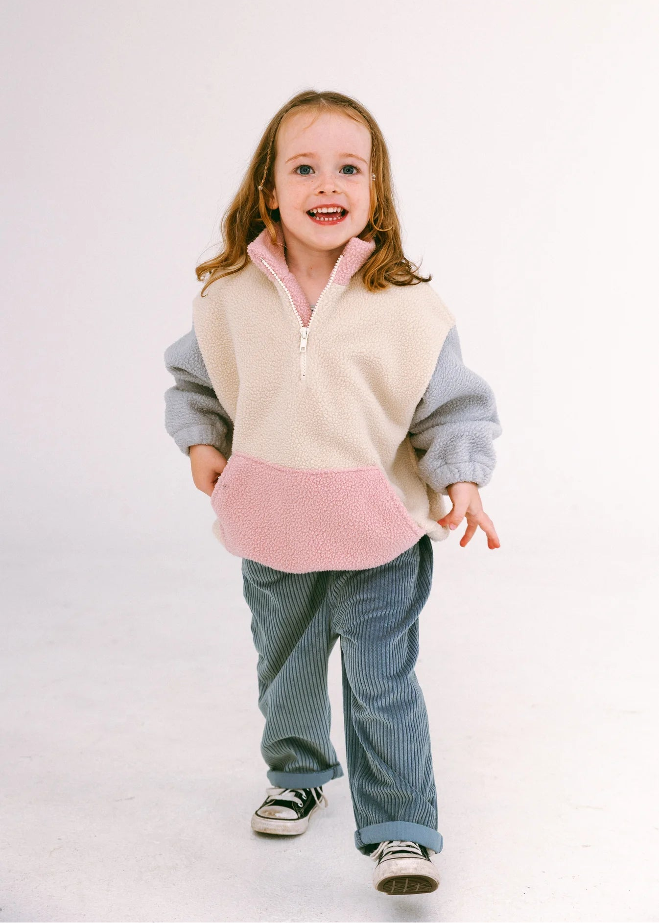 A young girl smiling and walking while wearing a color-blocked quarter-zip fleece pullover with a cream body, light gray sleeves, and a pink pocket, paired with blue corduroy pants and black-and-white sneakers, against a plain white studio background.
