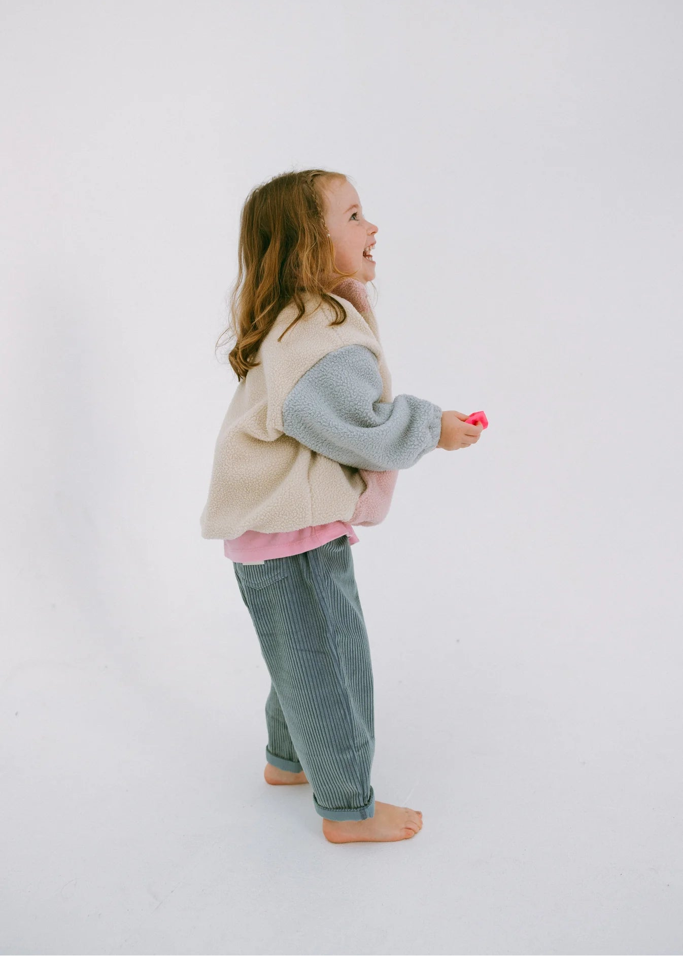 Young girl standing on a white background wearing a beige vest, gray sweater, pink shirt, and blue jeans.