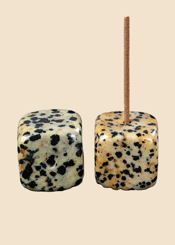 Two square polished stone beads with black and cream speckles, one standing upright with a tan string threaded through it, on a solid beige background.