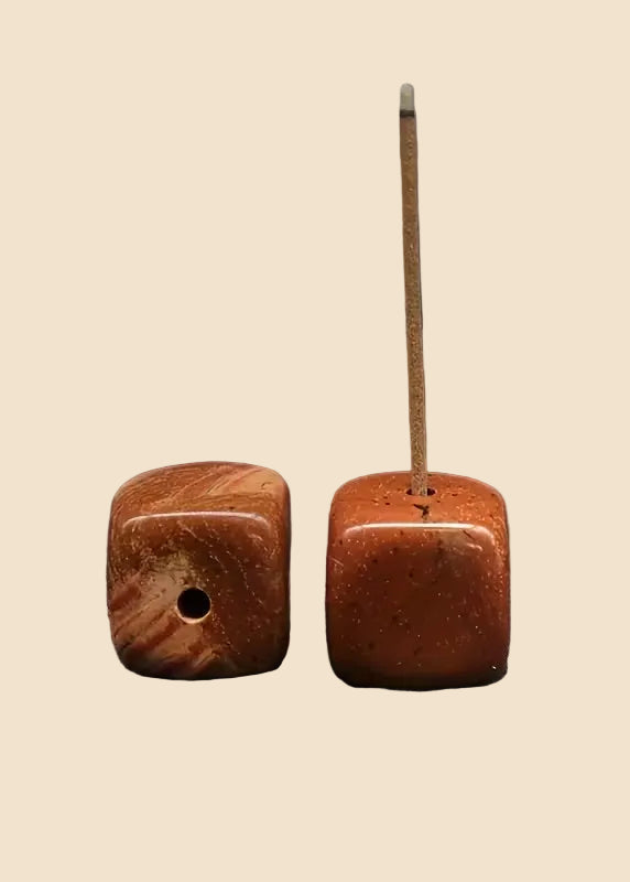 Two square brown stone beads with a hole, one standing upright with a thin incense stick inserted, on a solid beige background.