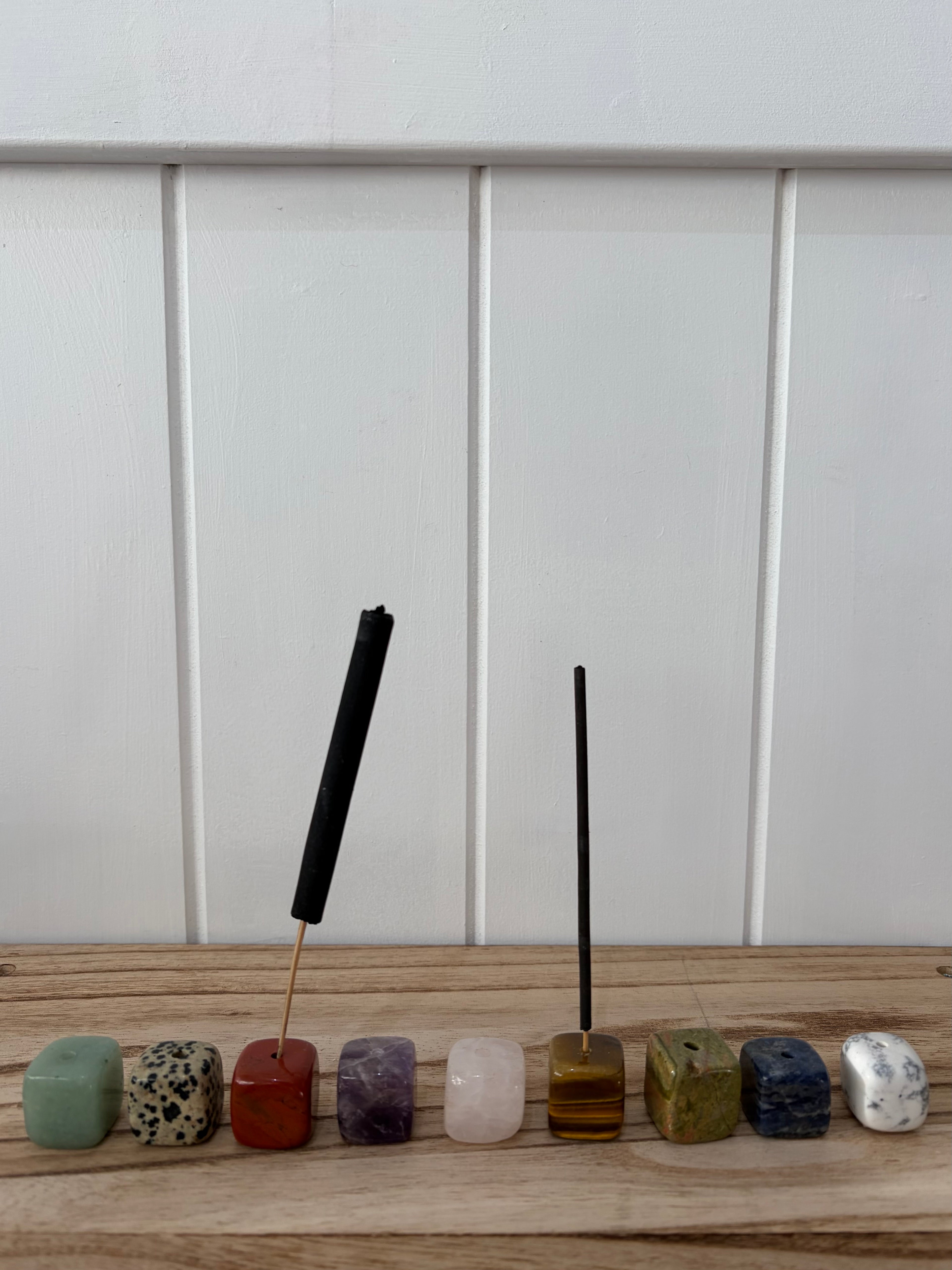 Two black incense sticks resting in square multicolored crystal holders lined up on a wooden surface against a white paneled wall.