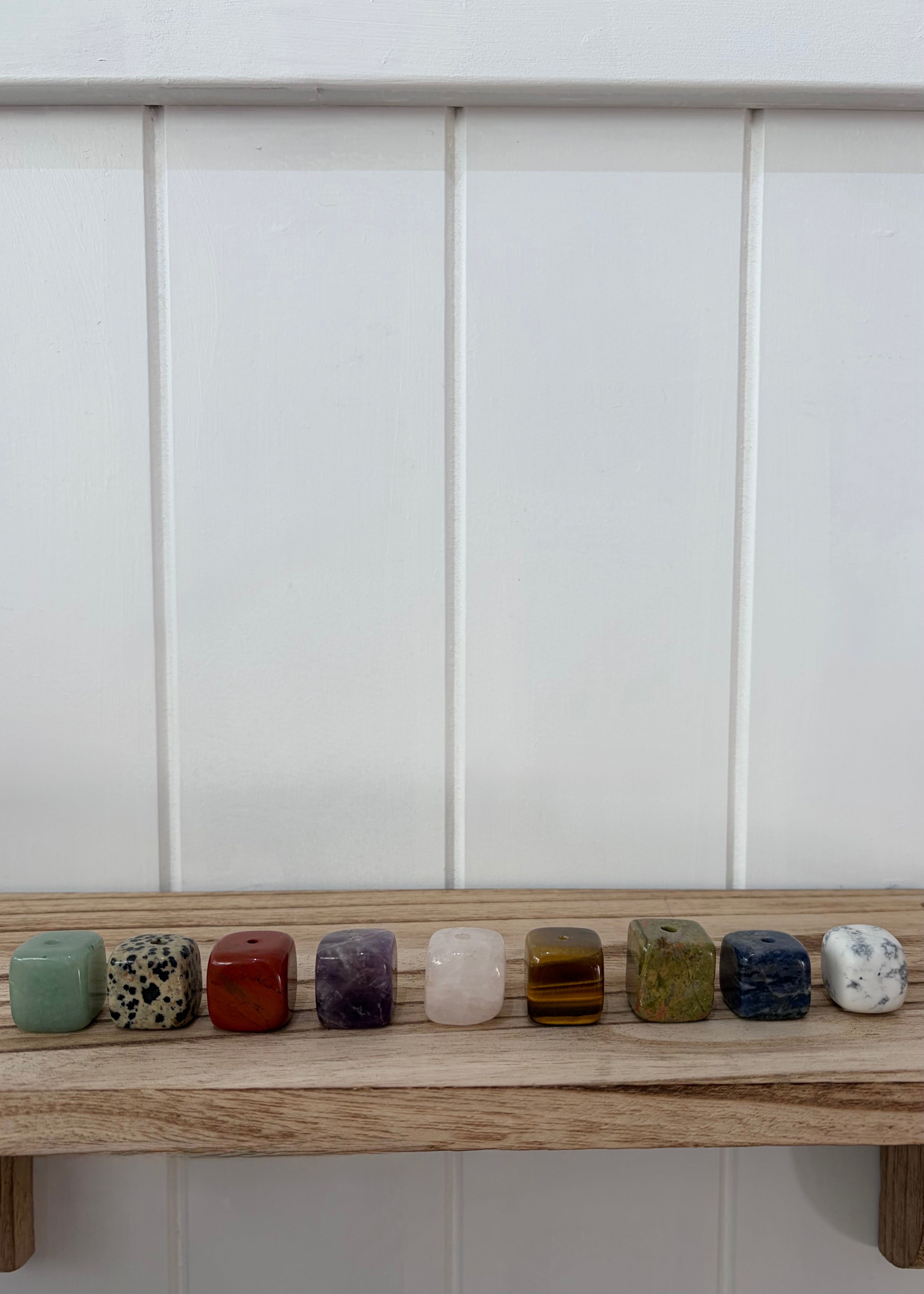 Nine small cylindrical stone beads or cabochons in assorted colors and patterns are lined up on a light wooden shelf against a white paneled wall.