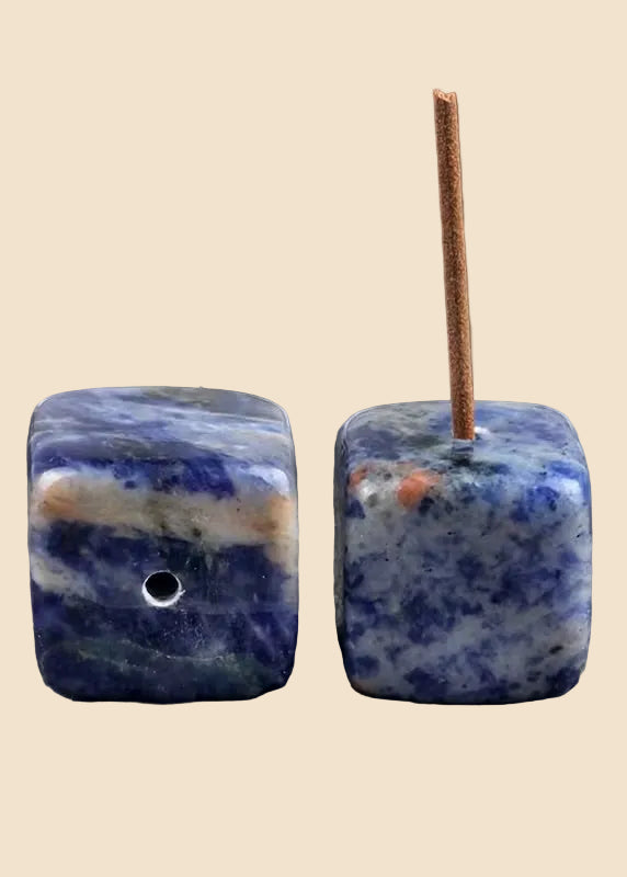 Two square blue lapis lazuli stone beads with a hole, shown from different angles on a solid beige background. One bead is oriented to show its blue and white marbled texture, and the other is shown from the side with a wooden incense stick inserted.