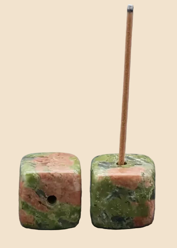 Two square polished stone beads with a pink and green marbled pattern, shown on a plain beige background. One bead has a hole on the side, and the other has a thin wooden incense stick inserted through the top.
