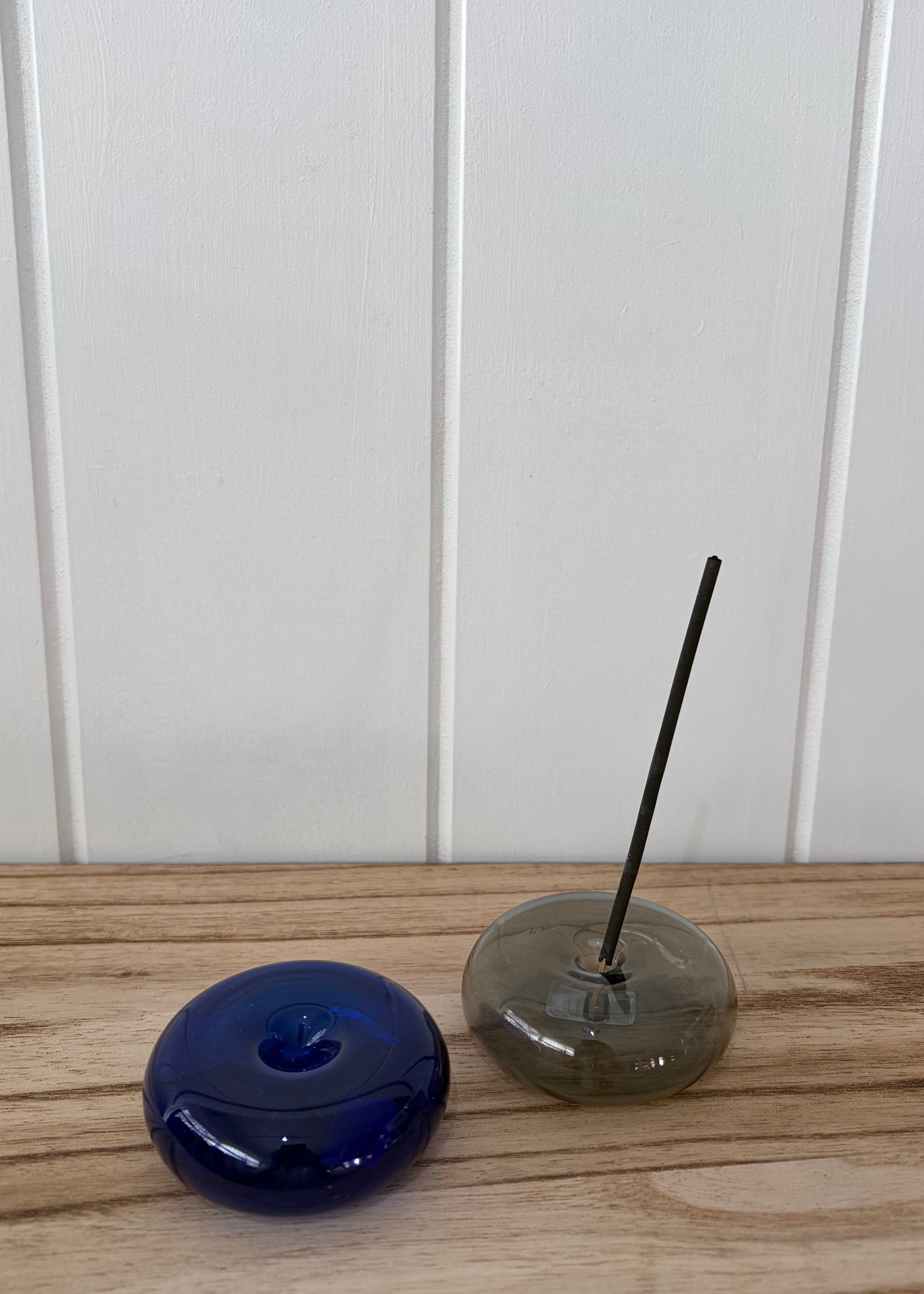 A dark blue glass incense holder with a matching blue stone top sits next to a smoky gray glass incense burner with a black incense stick, displayed on a light wood surface against a white paneled wall.