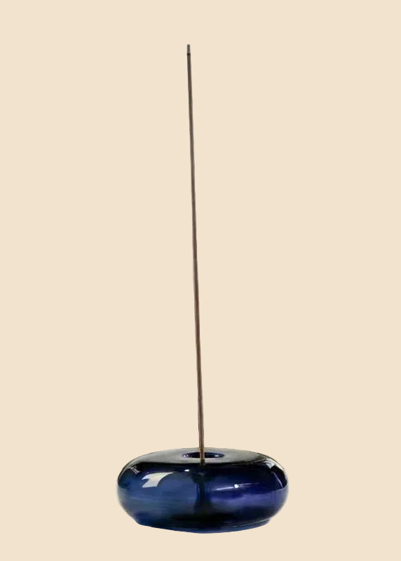 A dark blue glass incense holder with a single incense stick inserted vertically, set against a solid light beige background.