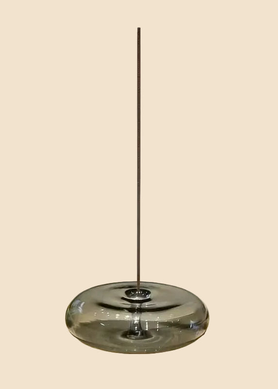 A modern pendant light with a thick, rounded translucent smoky glass shade and a thin vertical metal rod extending upwards, shown against a solid light beige background.