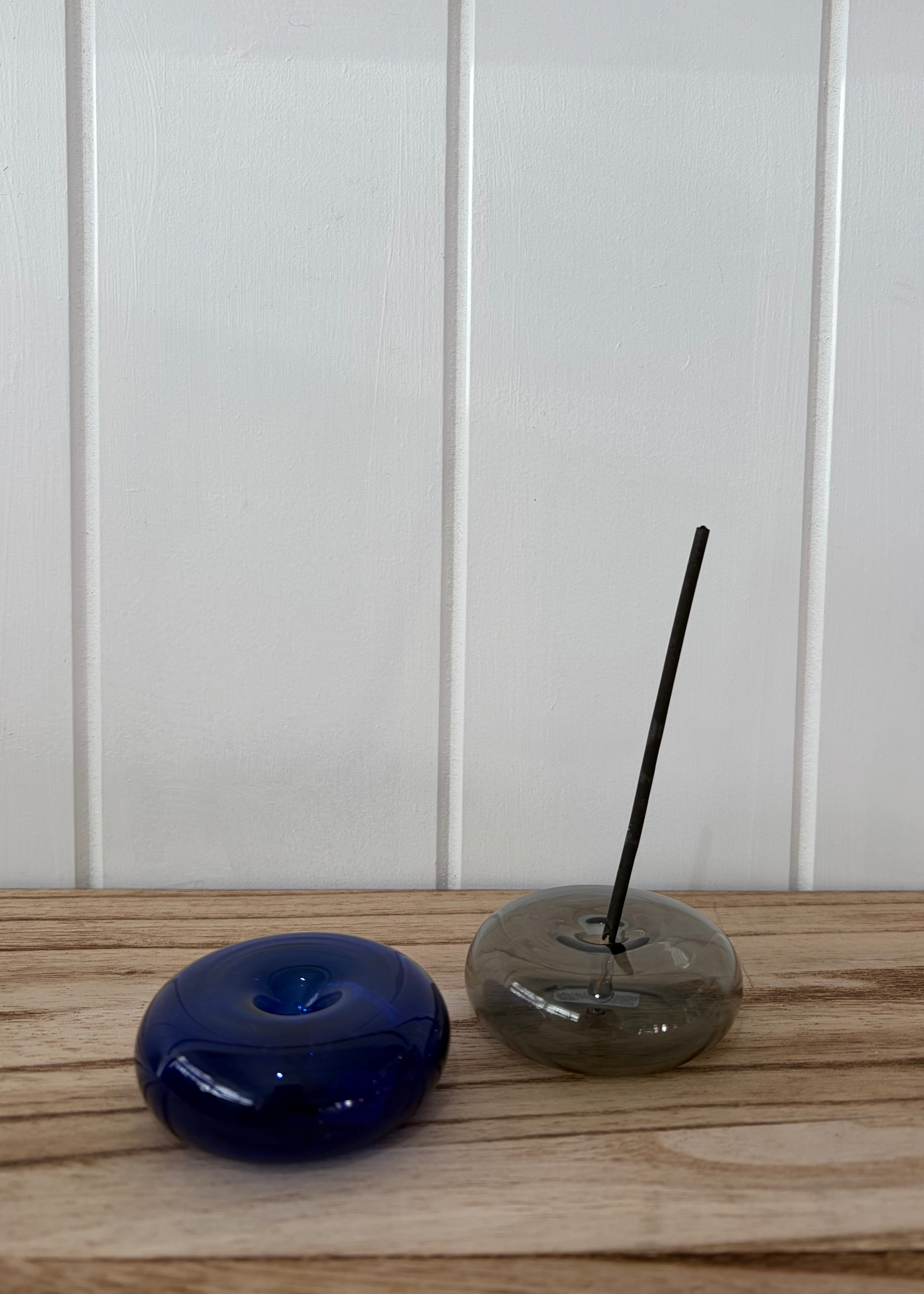 A dark blue glass donut-shaped incense holder and a smoky gray glass donut-shaped incense holder with a black incense stick, placed on a light wooden surface against a white paneled wall.
