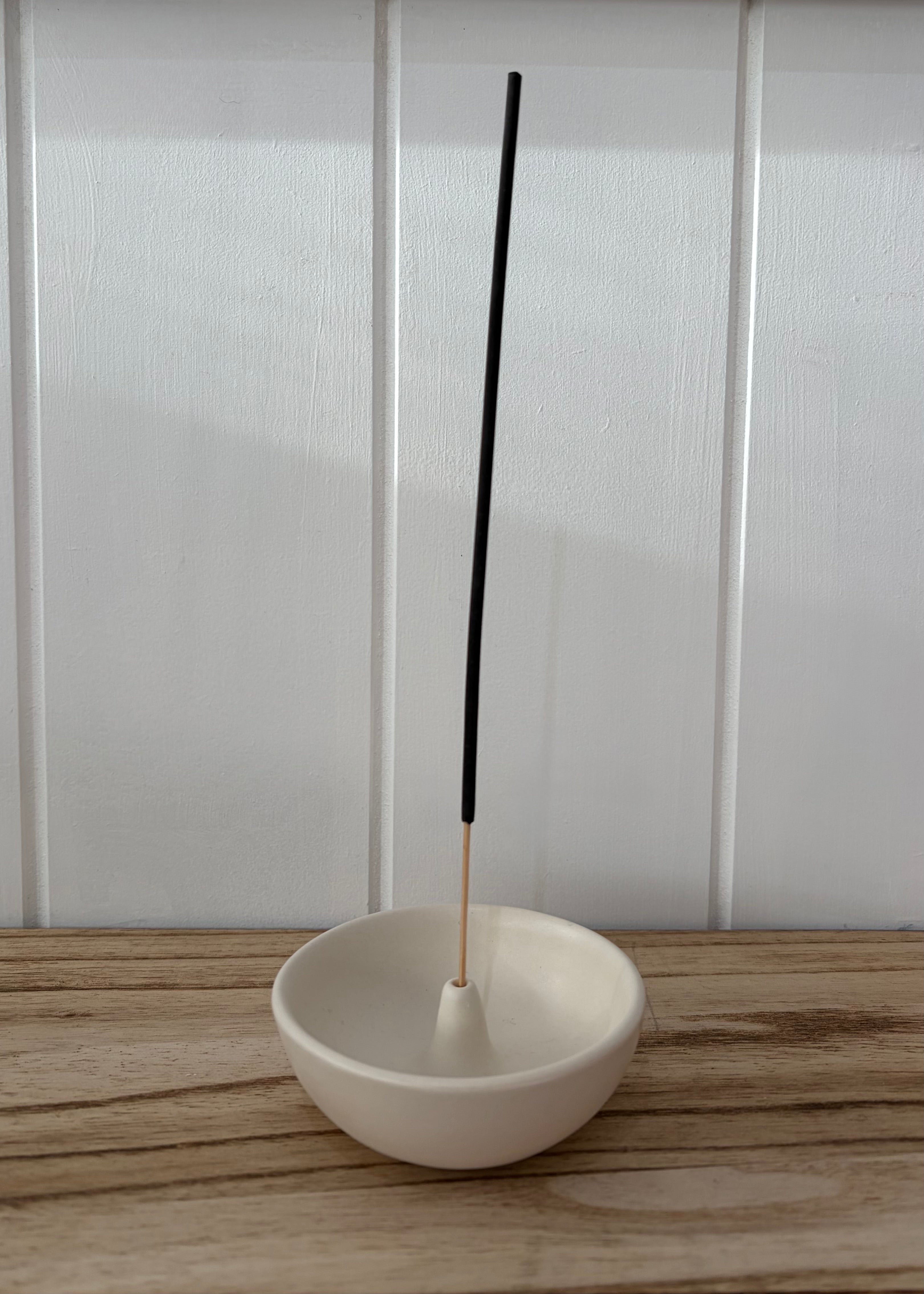 A white ceramic incense holder bowl with a single black incense stick standing upright inside, placed on a light wood surface against a white paneled wall.
