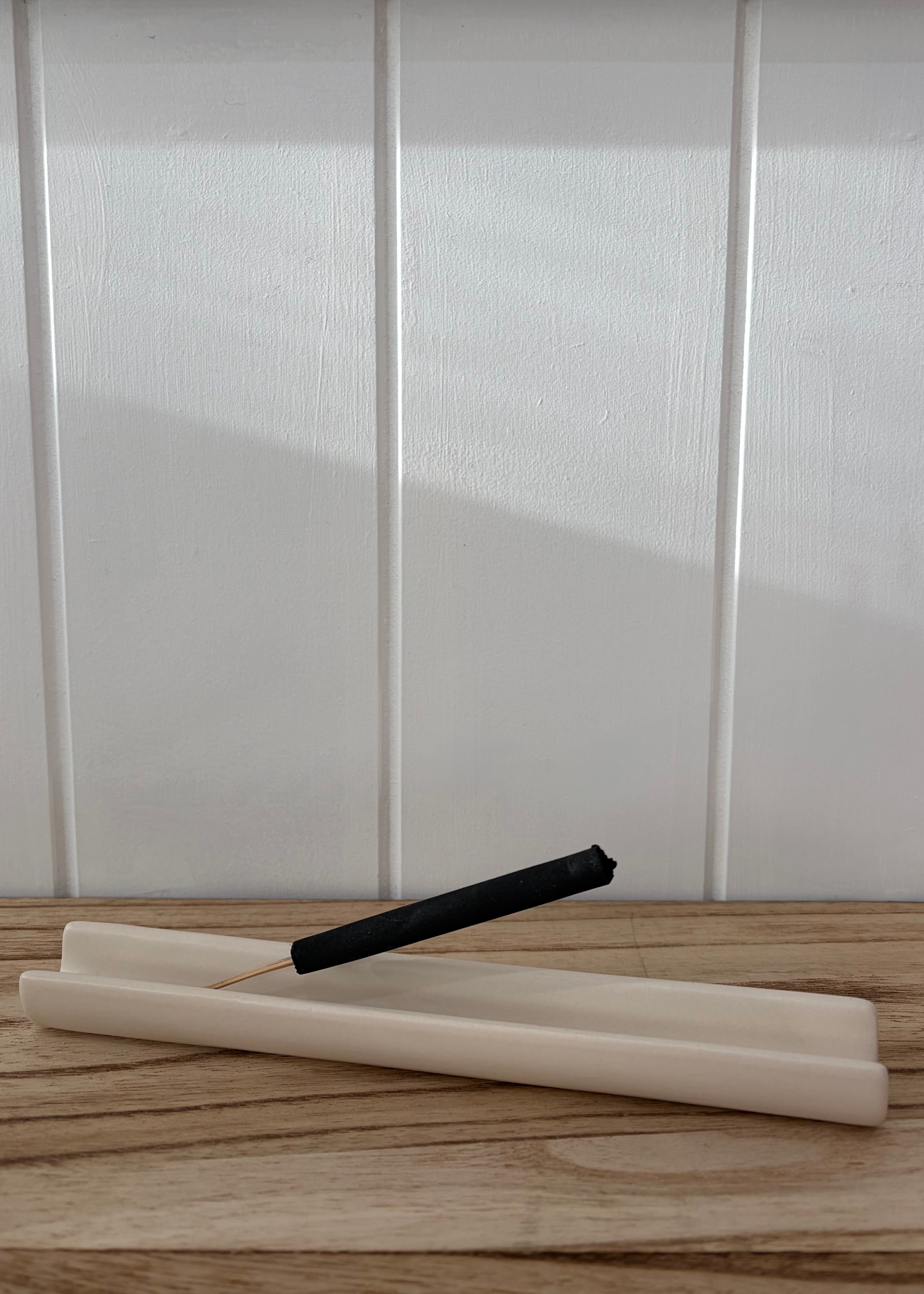 A black incense stick resting on two cream-colored wax candles on a light wood surface, set against a white paneled wall.
