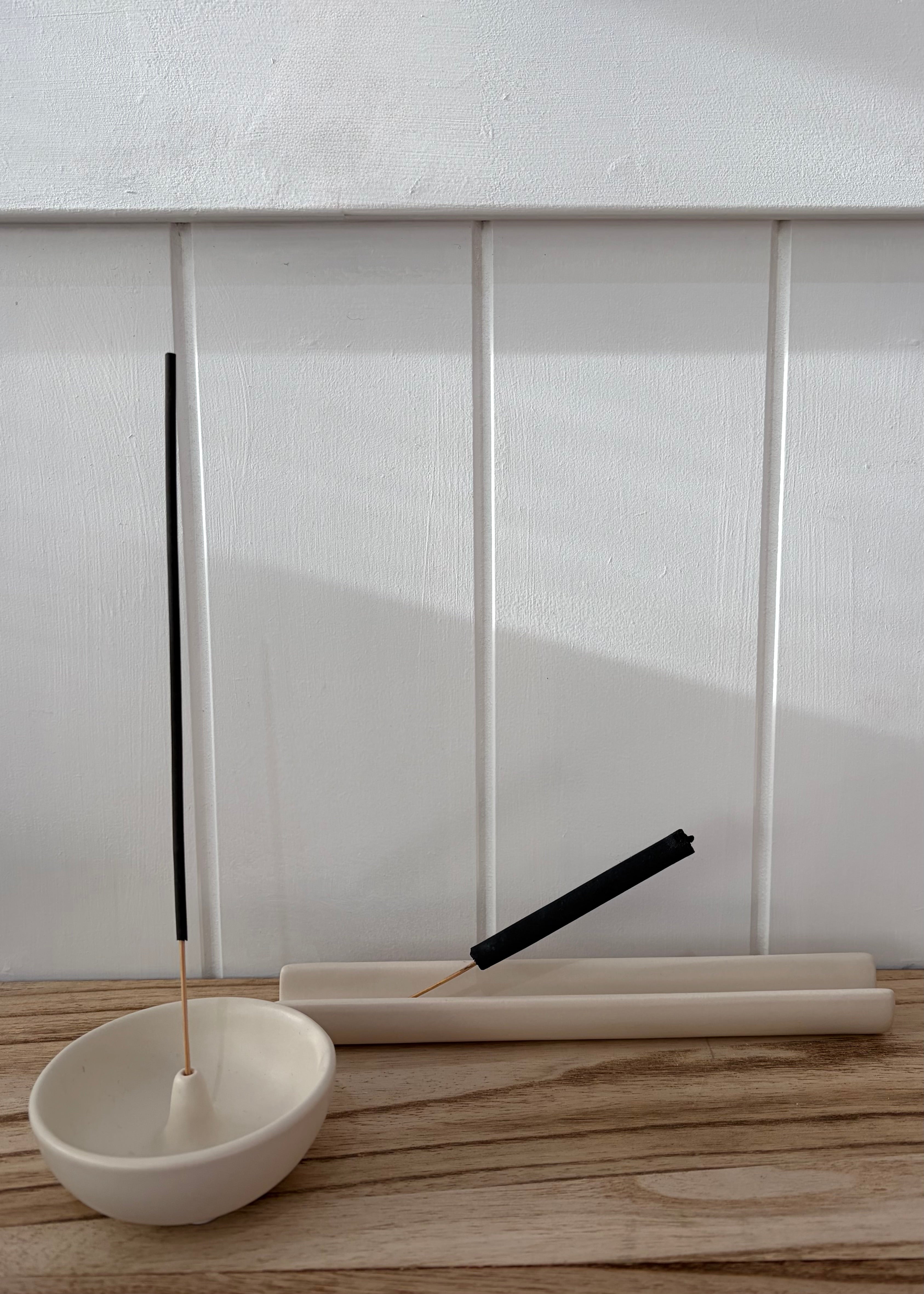 Minimalist incense holder set featuring a small white ceramic bowl with a black incense stick, a matching white cylindrical holder with another black incense stick resting diagonally, and a light wood tabletop against a white paneled wall.