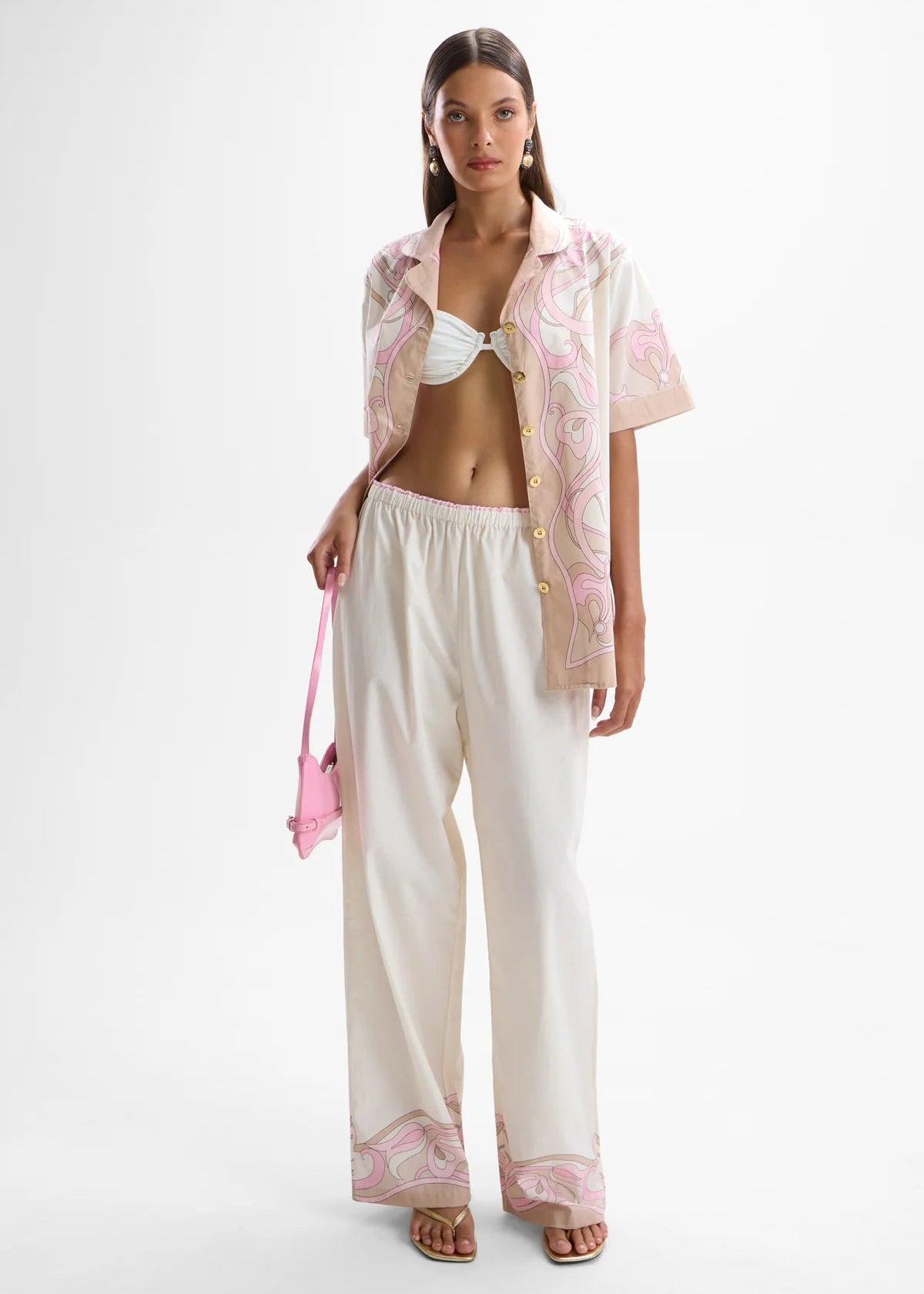 Woman wearing a pink floral blouse, white pants, and holding a pink bag on a white background