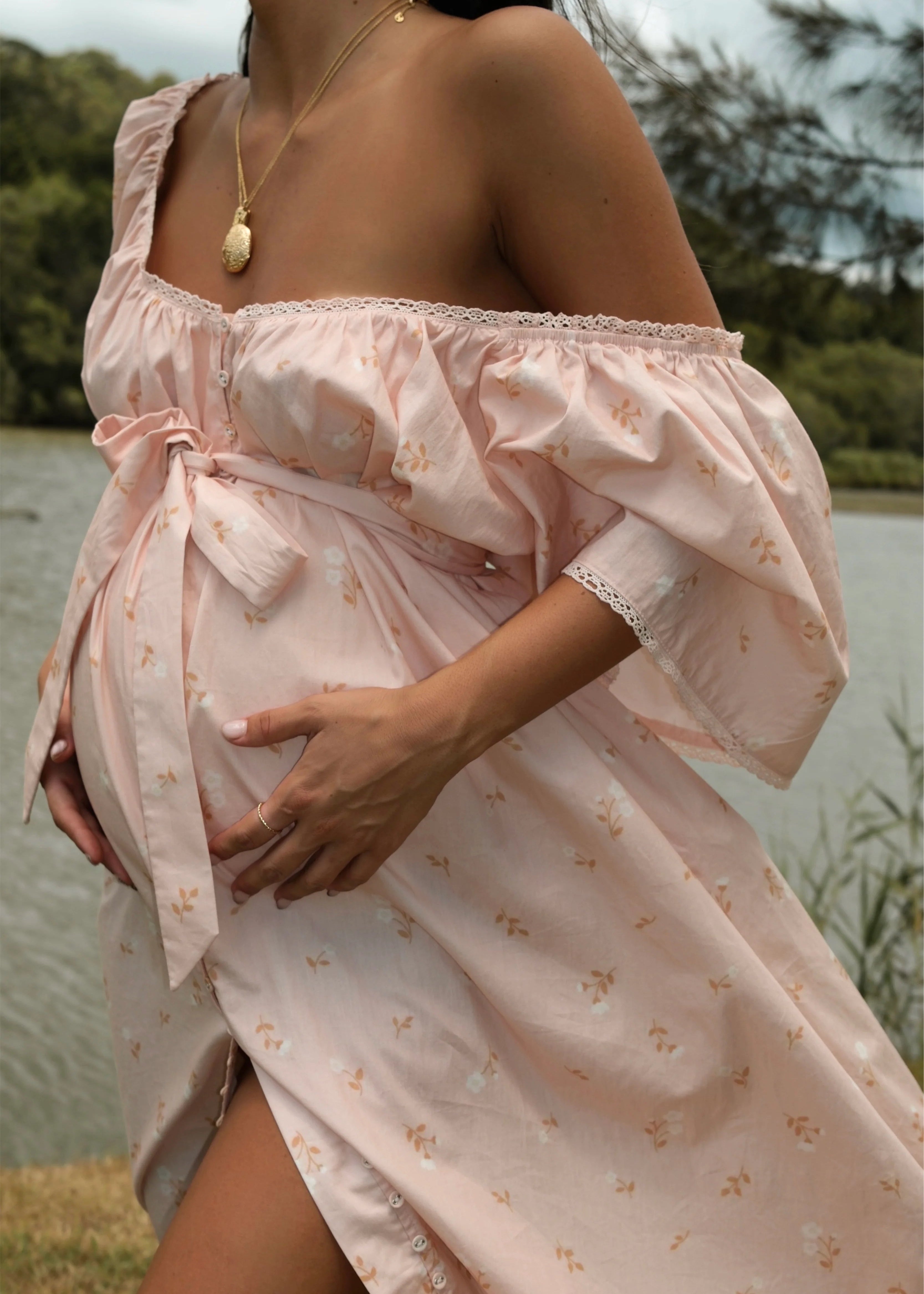 Woman wearing a pink off-shoulder dress with a floral pattern, standing by a body of water.