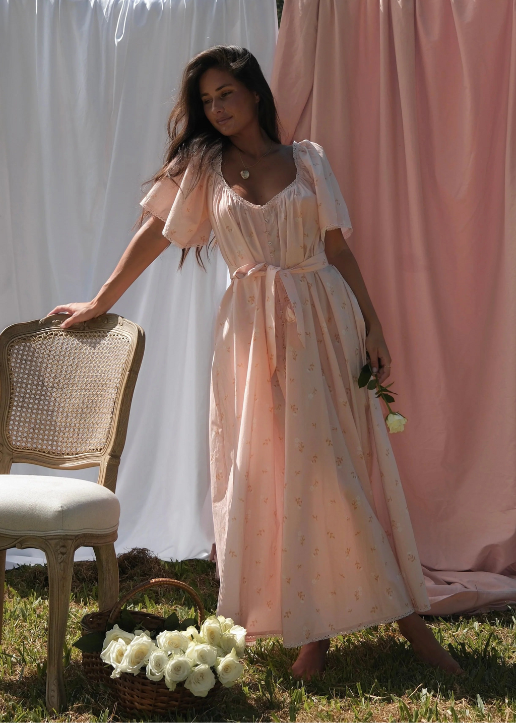 Woman in a peach dress standing next to a chair with white flowers, against a backdrop of white and pink fabric.