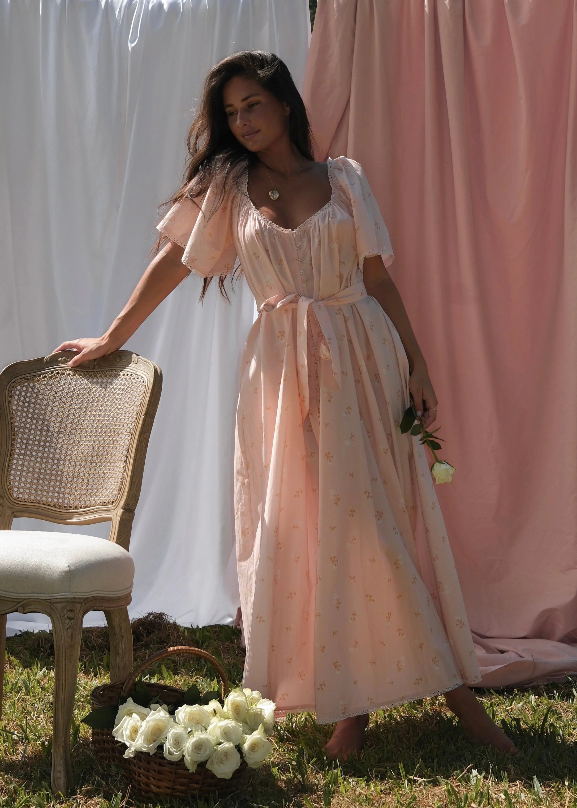 Woman in a peach dress standing next to a chair with white flowers, against a backdrop of white and pink fabric.
