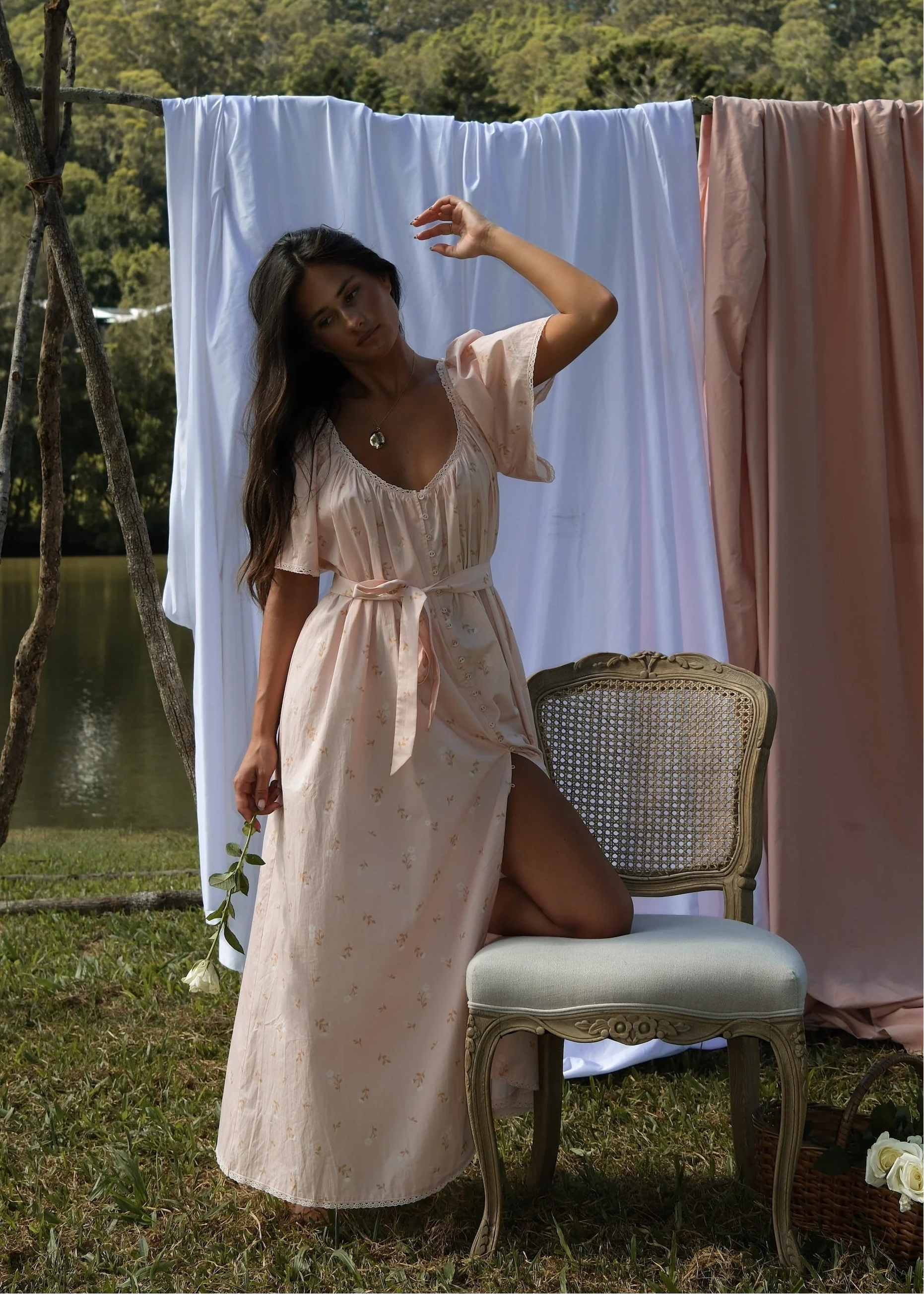 Woman in a flowing dress standing next to a chair with draped fabrics in an outdoor setting.