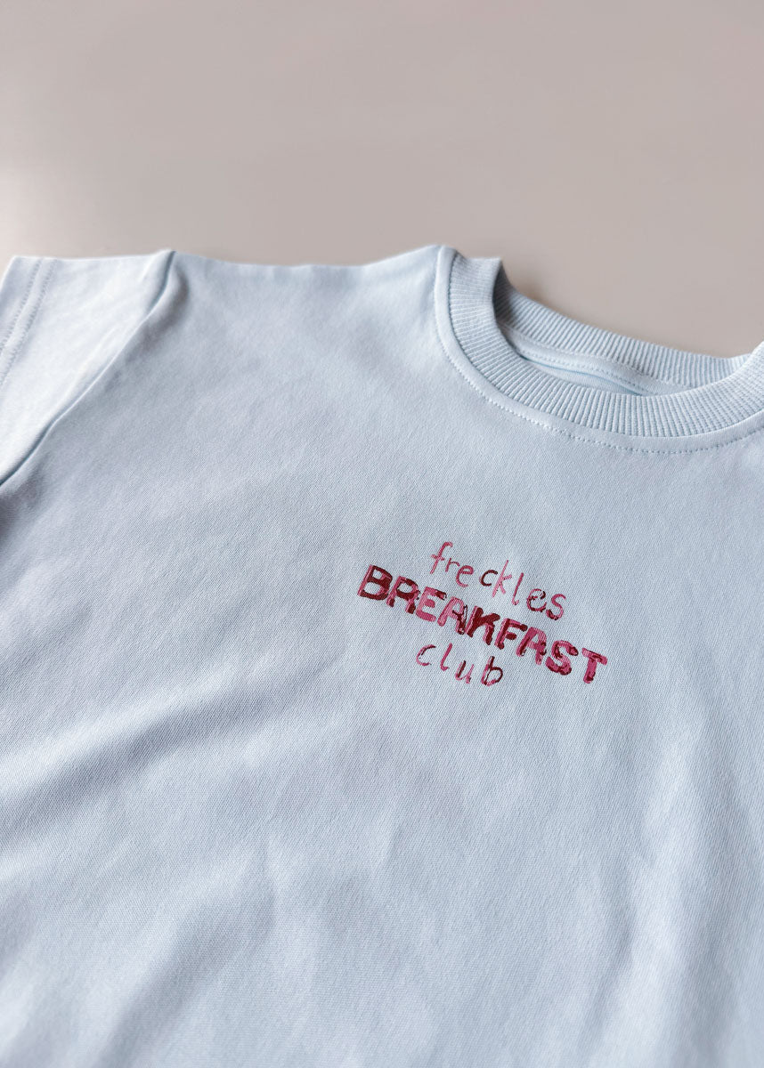 White sweatshirt with red text 'freckles BREAKFAST club' on a light gray background