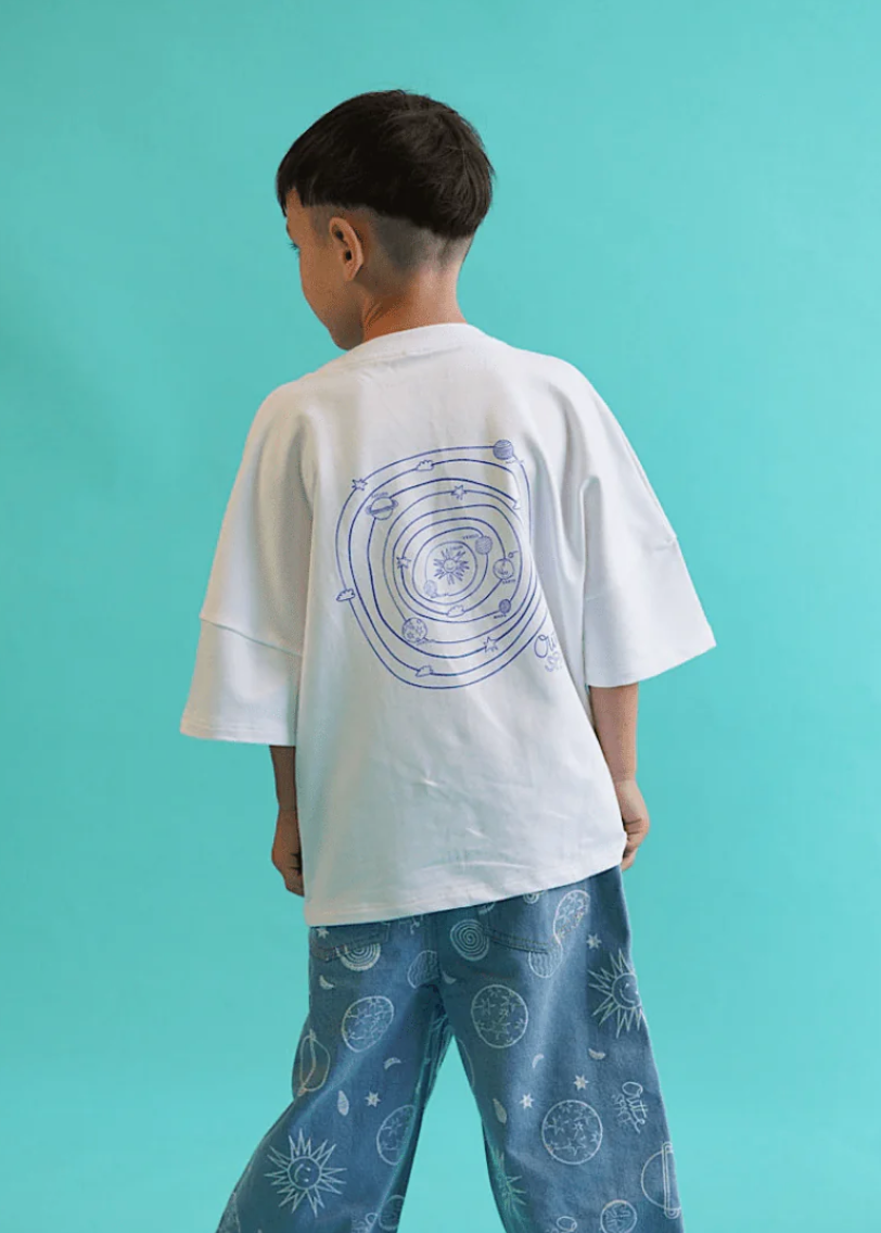 Child wearing a white t-shirt with a design and blue pants with planet patterns against a teal background