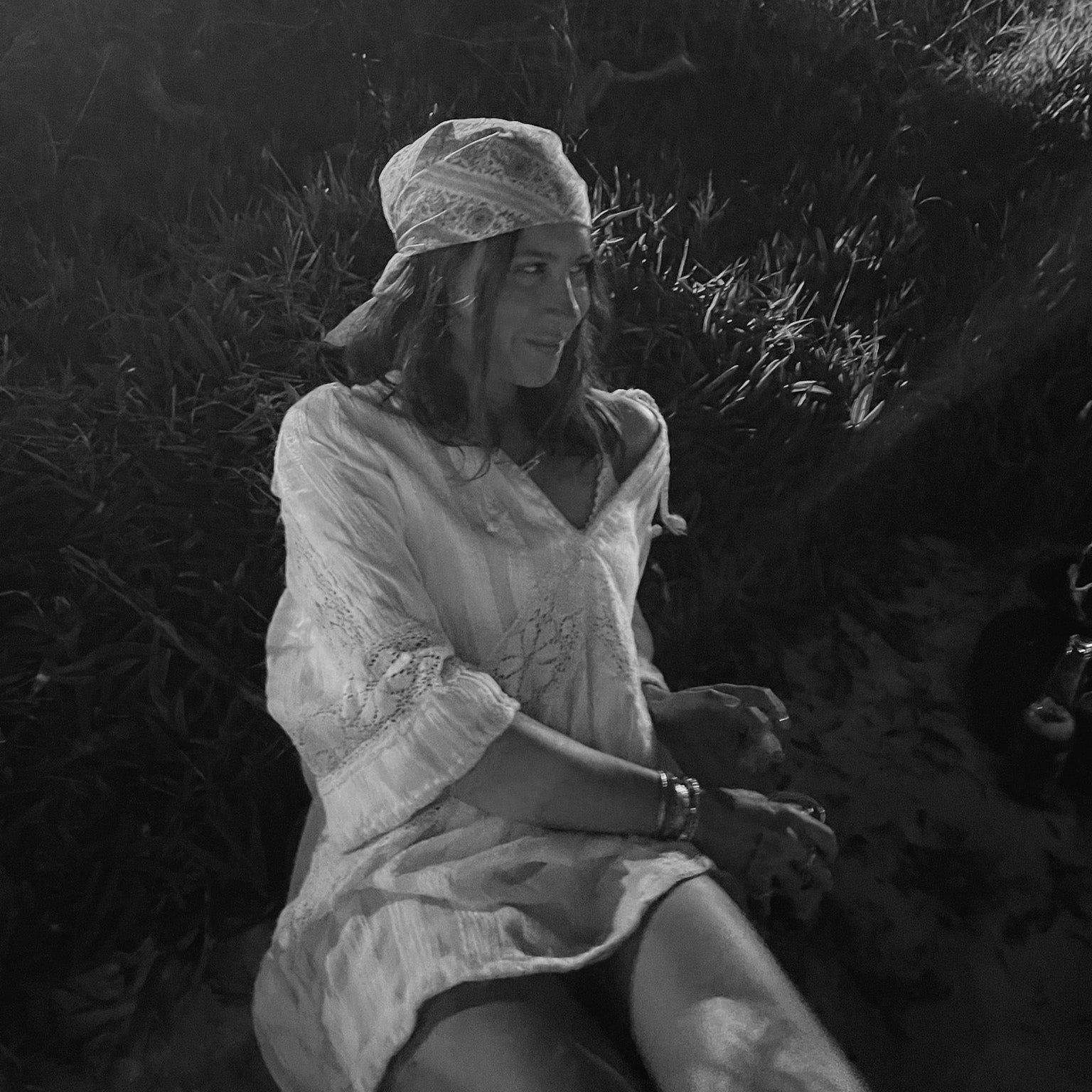 A woman sitting outdoors in a grassy area at night, wearing a light-colored, loose-fitting dress and a matching headscarf. The photo is in black and white, with dark shadows and tall grass in the background.