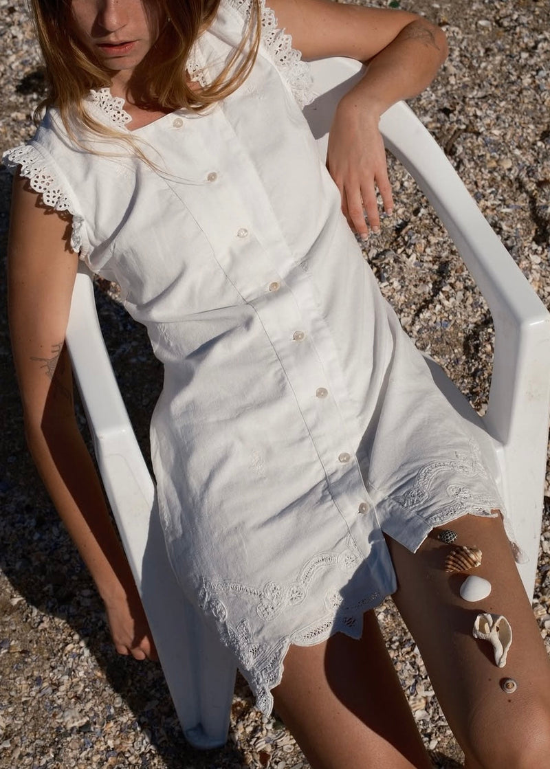 Woman in a white dress sitting on a white chair on a pebbly beach.