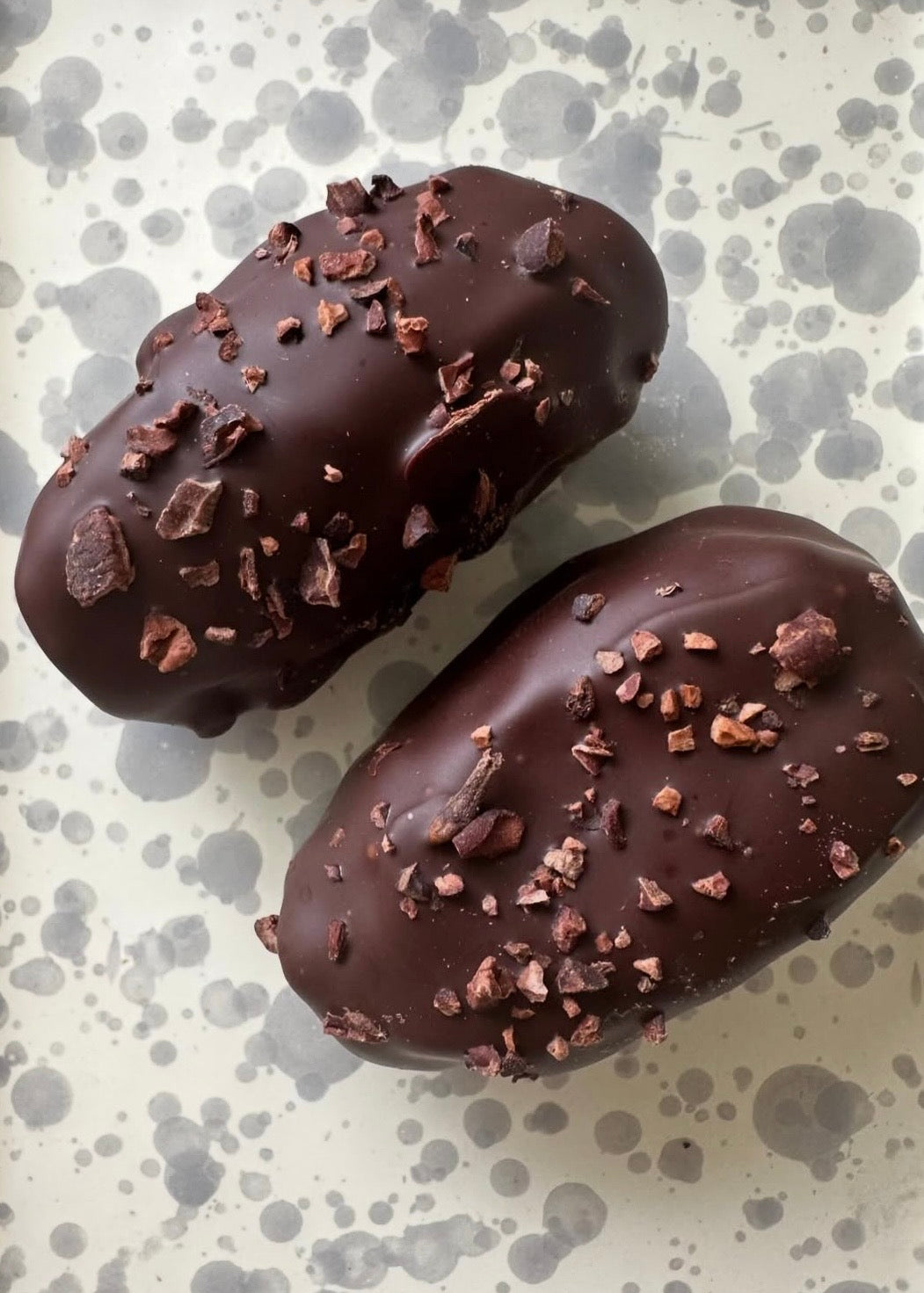 Two chocolate-covered treats with chocolate shavings on a speckled surface