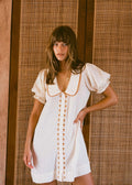 A woman wearing a short white dress with puff sleeves, a Peter Pan collar, and decorative gold buttons down the front, posing in front of a woven rattan screen.