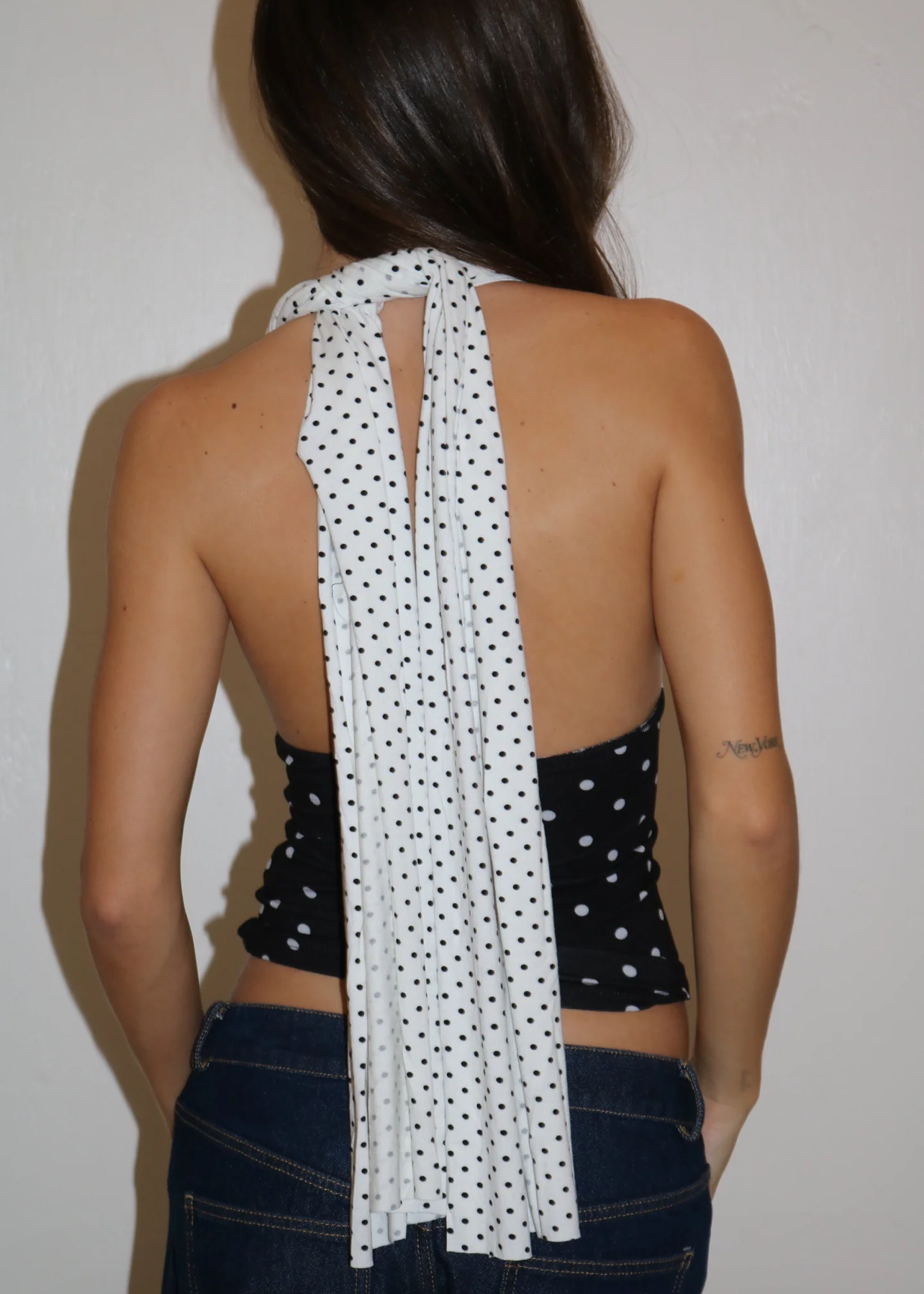 Person wearing a black halter top with white polka dots and a long white scarf over their shoulder, on a plain background.