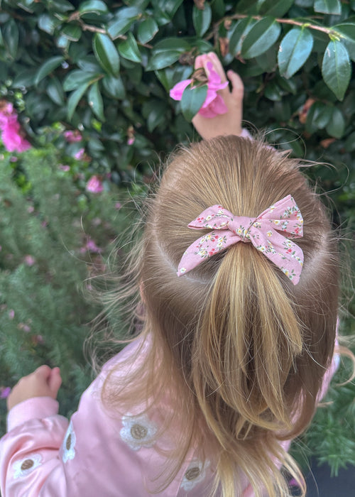 Child with blonde hair tied back with a pink bow, wearing a pink jacket with floral patterns, against a green bush background.