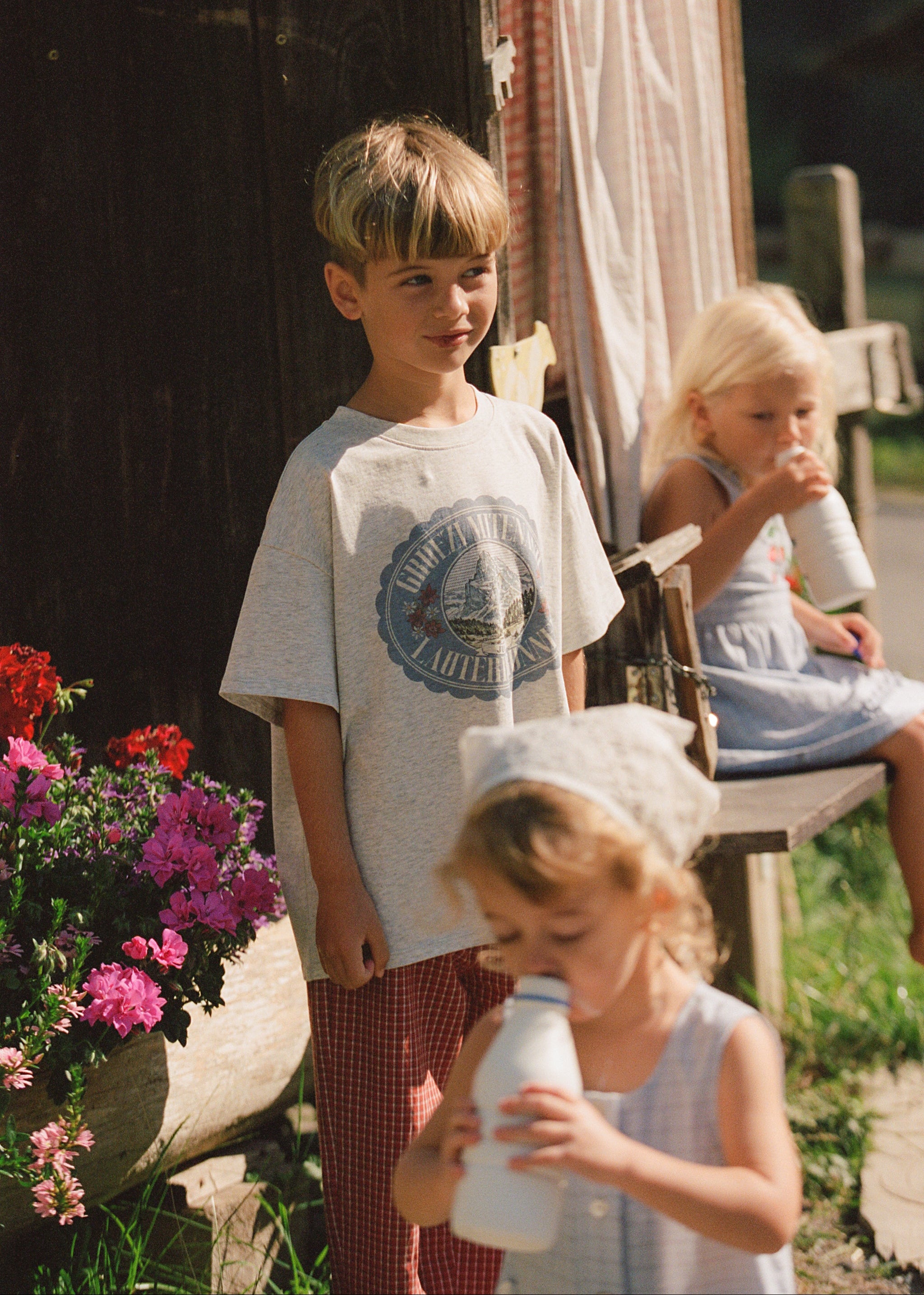 A young boy wearing a light gray graphic t-shirt and red gingham pants stands next to a rustic wooden porch, while two young girls in light blue dresses drink milk from white bottles. Pink flowers and a rustic wooden structure are visible in the background.