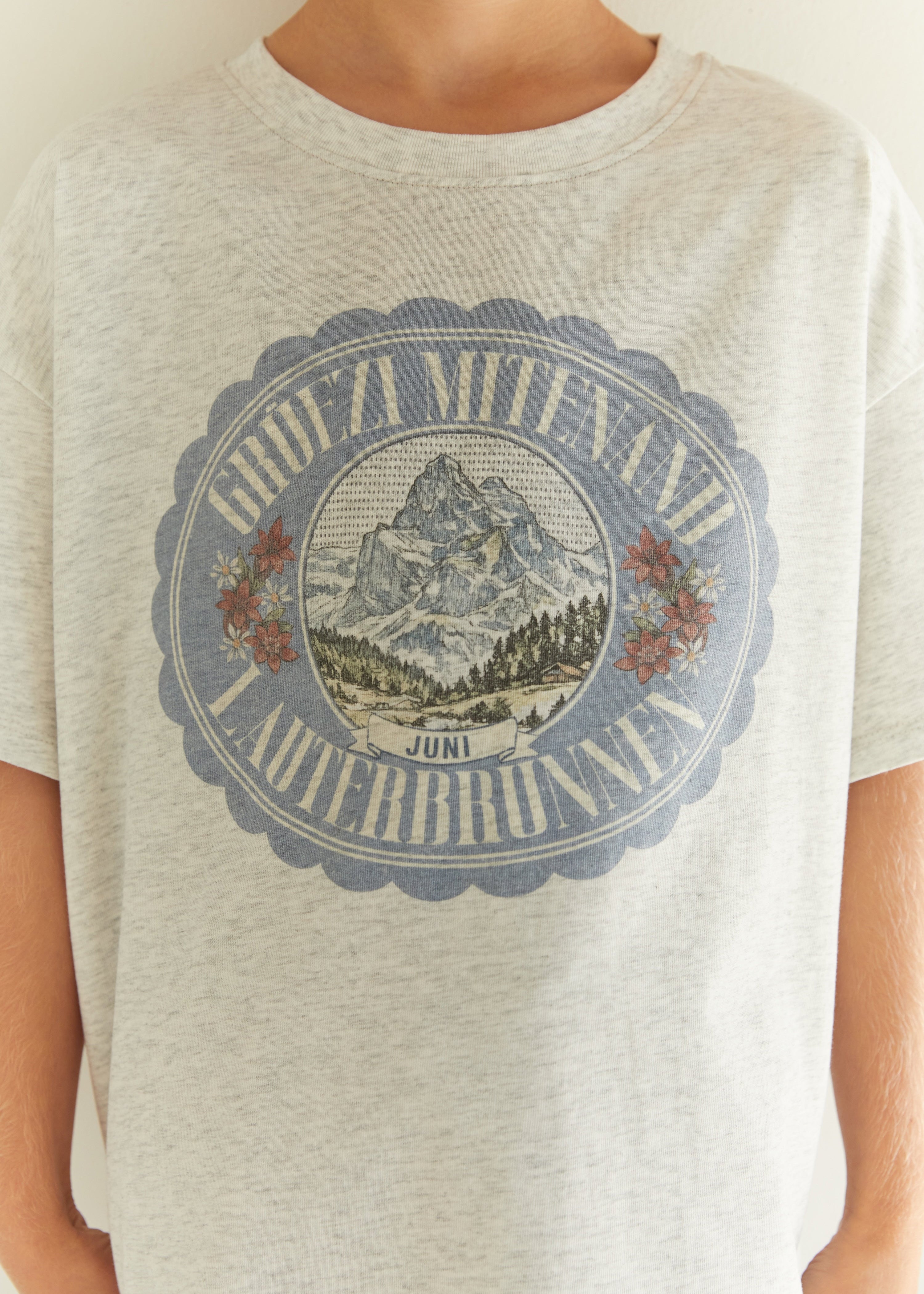 A close-up of a person wearing a light heather gray t-shirt with a vintage-style circular graphic on the chest featuring a mountain illustration and the text 'AUBRA Mitenadi Auberbronne Juni'.