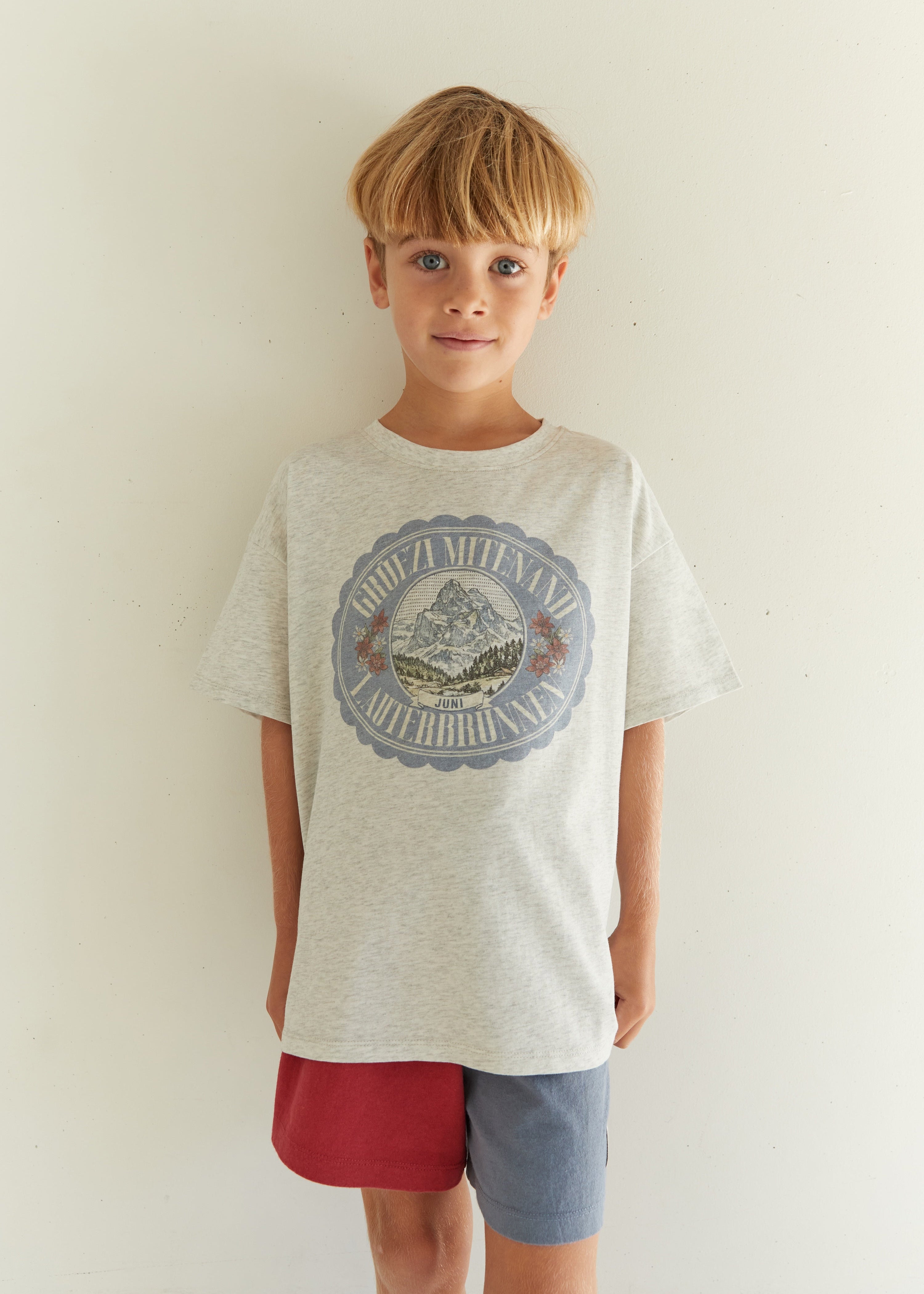 A young boy with blonde hair stands against a plain off-white wall wearing a light heather gray graphic t-shirt and a split red and blue shorts.
