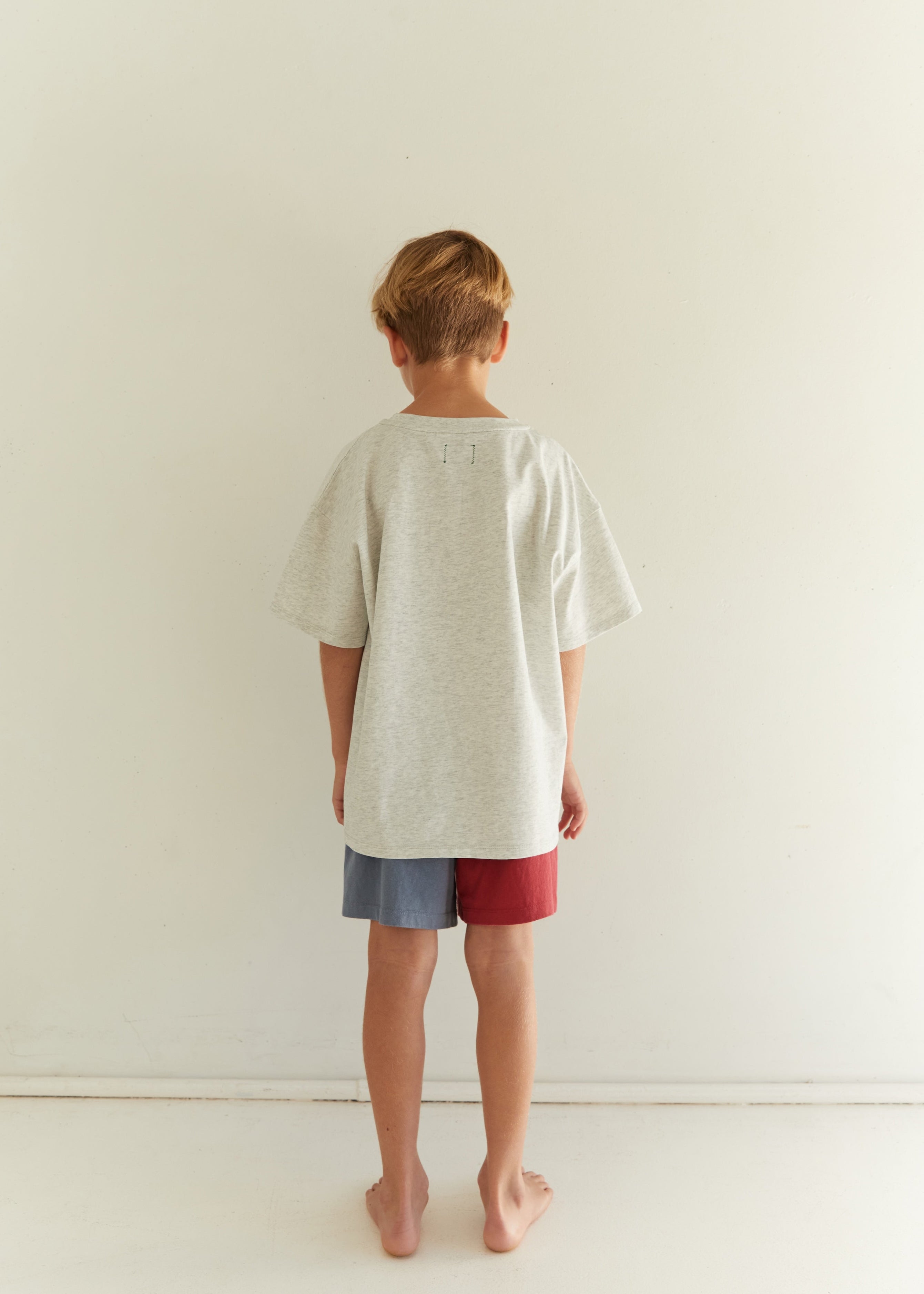 A child standing with their back to the camera, wearing an oversized light gray t-shirt and shorts with blue and red side panels, in a minimalist white room.
