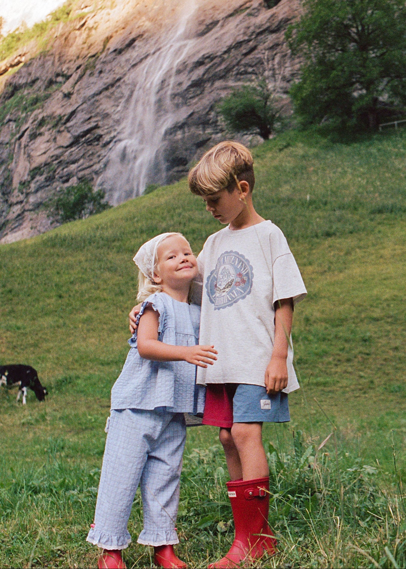 Two children standing in a grassy mountain meadow with a waterfall and rocky cliff in the background. The child on the left wears a light blue outfit and white headscarf, while the child on the right wears a white graphic T-shirt, blue shorts, and bright red rain boots.