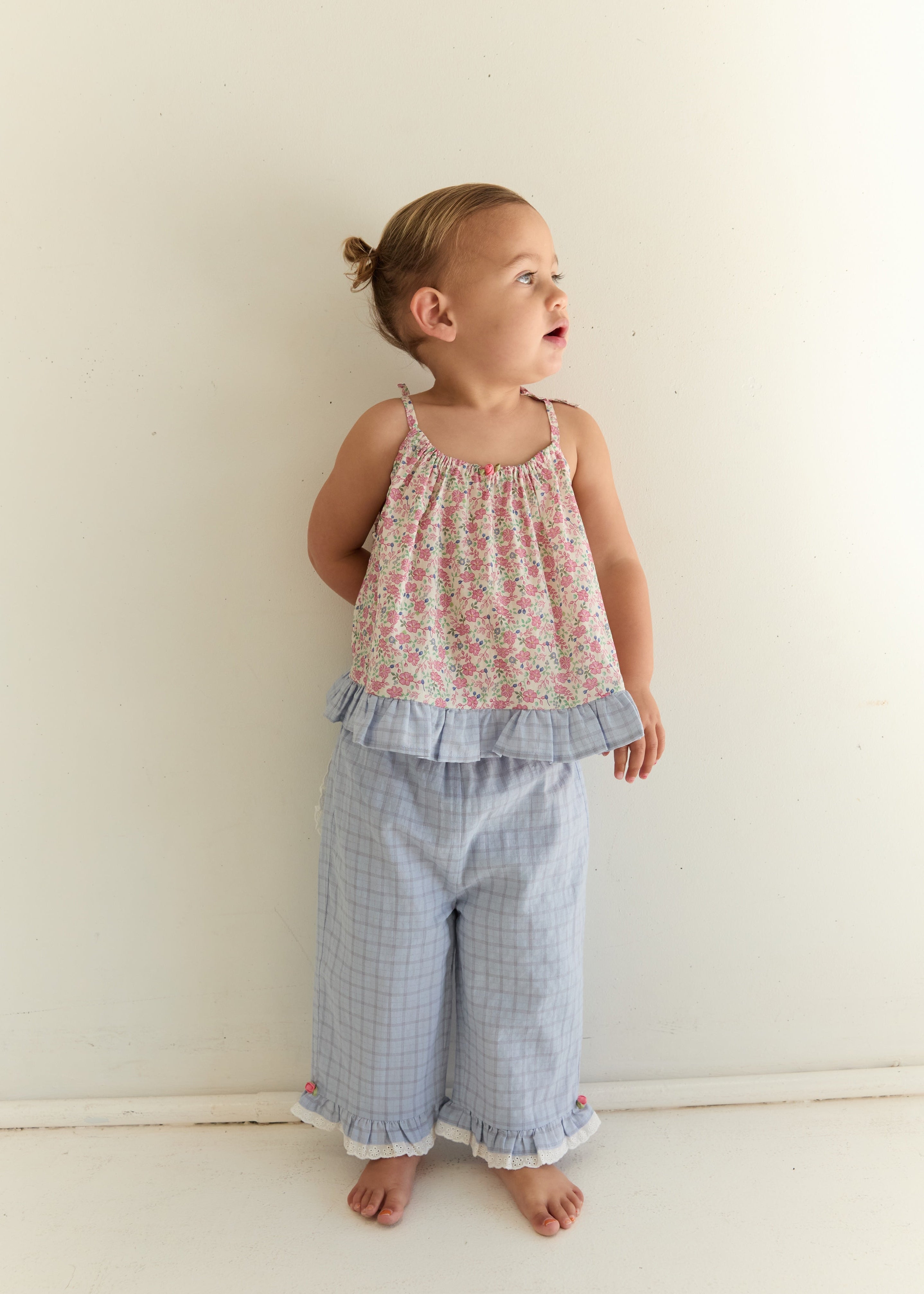 A toddler stands against a plain off-white wall wearing a two-piece outfit with a floral print halter top and light blue plaid pants with ruffled trim.