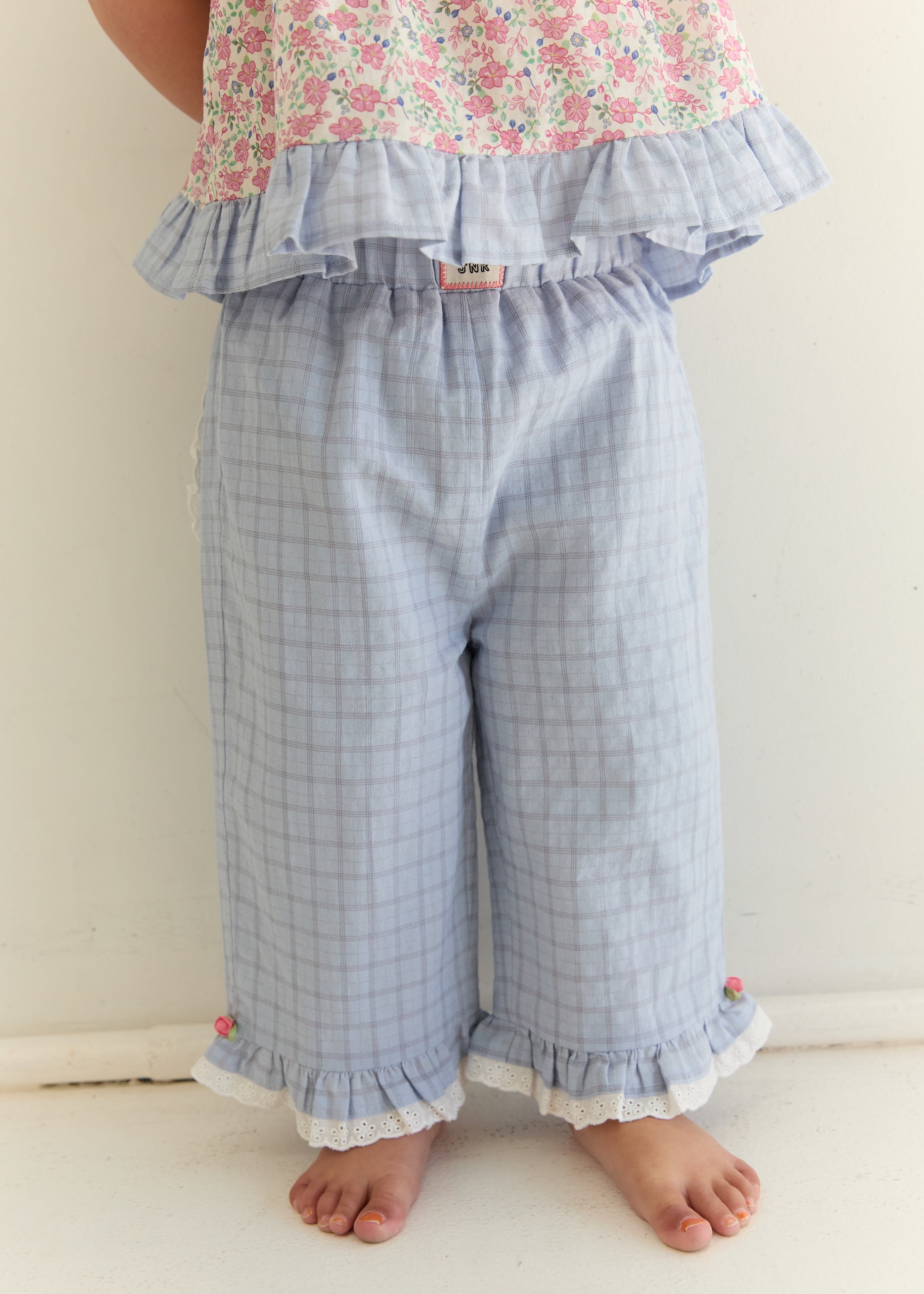 A child wearing light blue and white plaid cropped pants with ruffled hems, paired with a pink floral top, standing barefoot against a plain off-white wall.