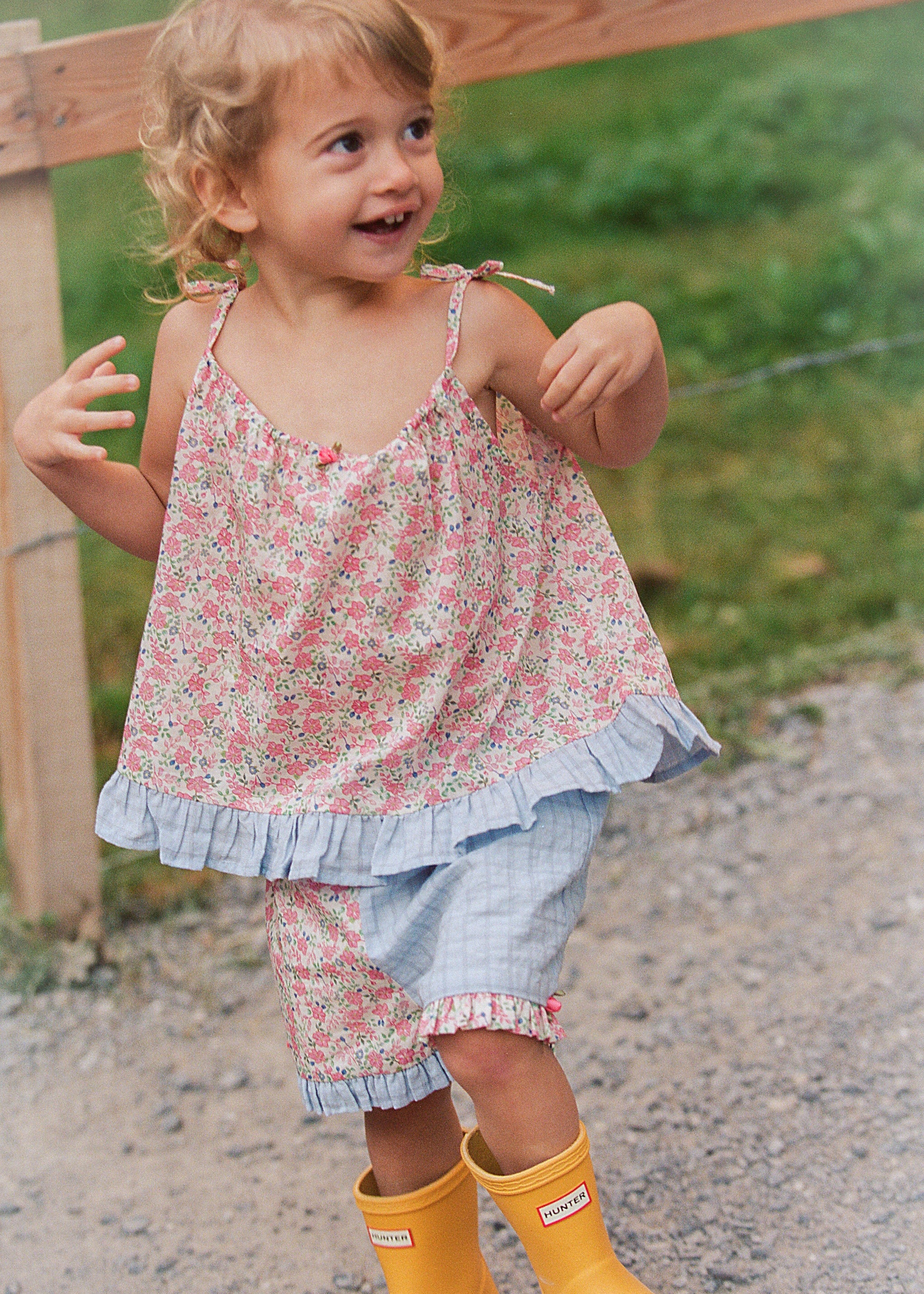 A young girl smiling outdoors, wearing a pink and white floral sleeveless top with a ruffled hem, matching floral shorts, and bright yellow rain boots.