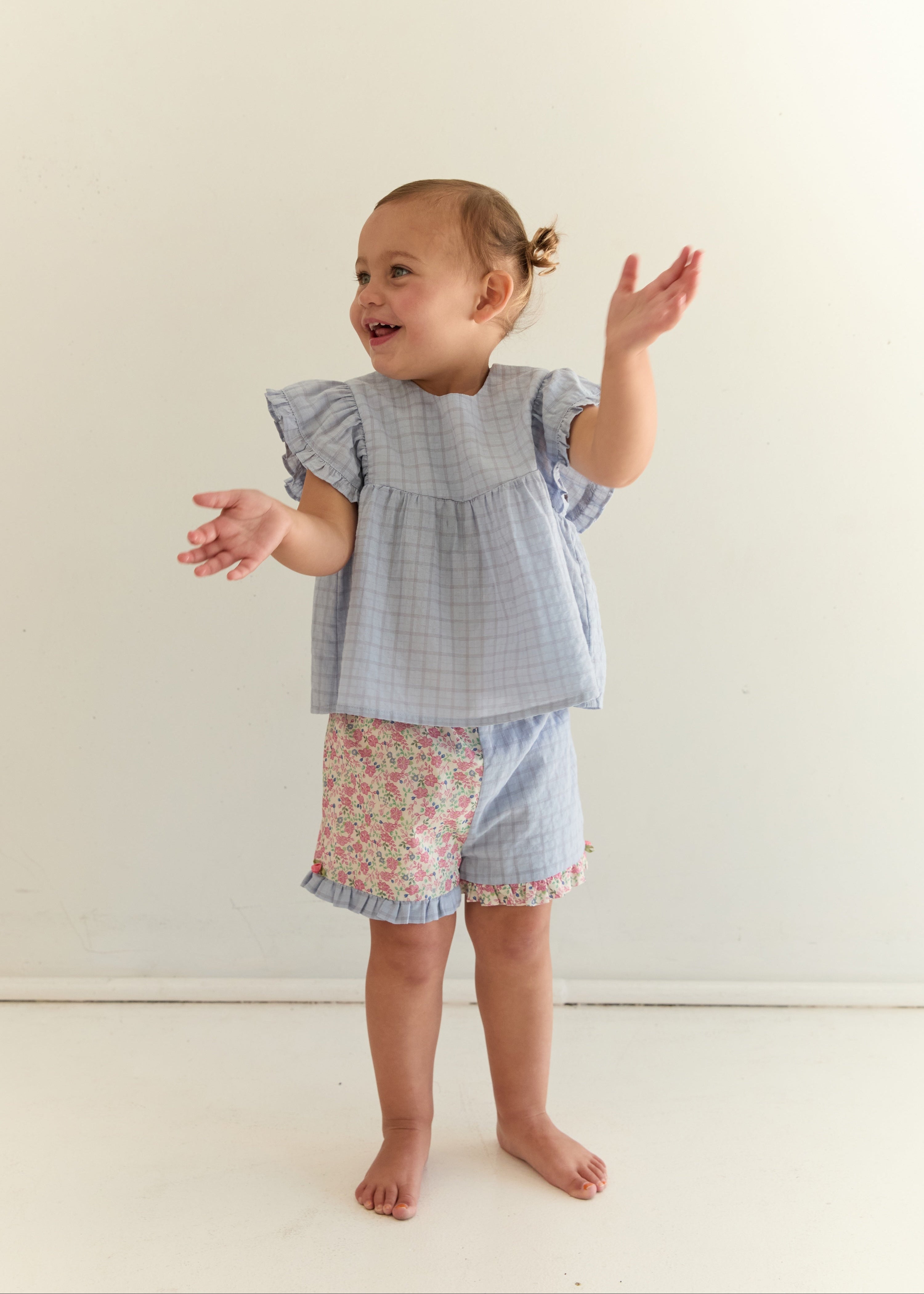 A toddler girl wearing a light blue and white gingham check romper with ruffled sleeves and floral print shorts, standing against a plain off-white background.