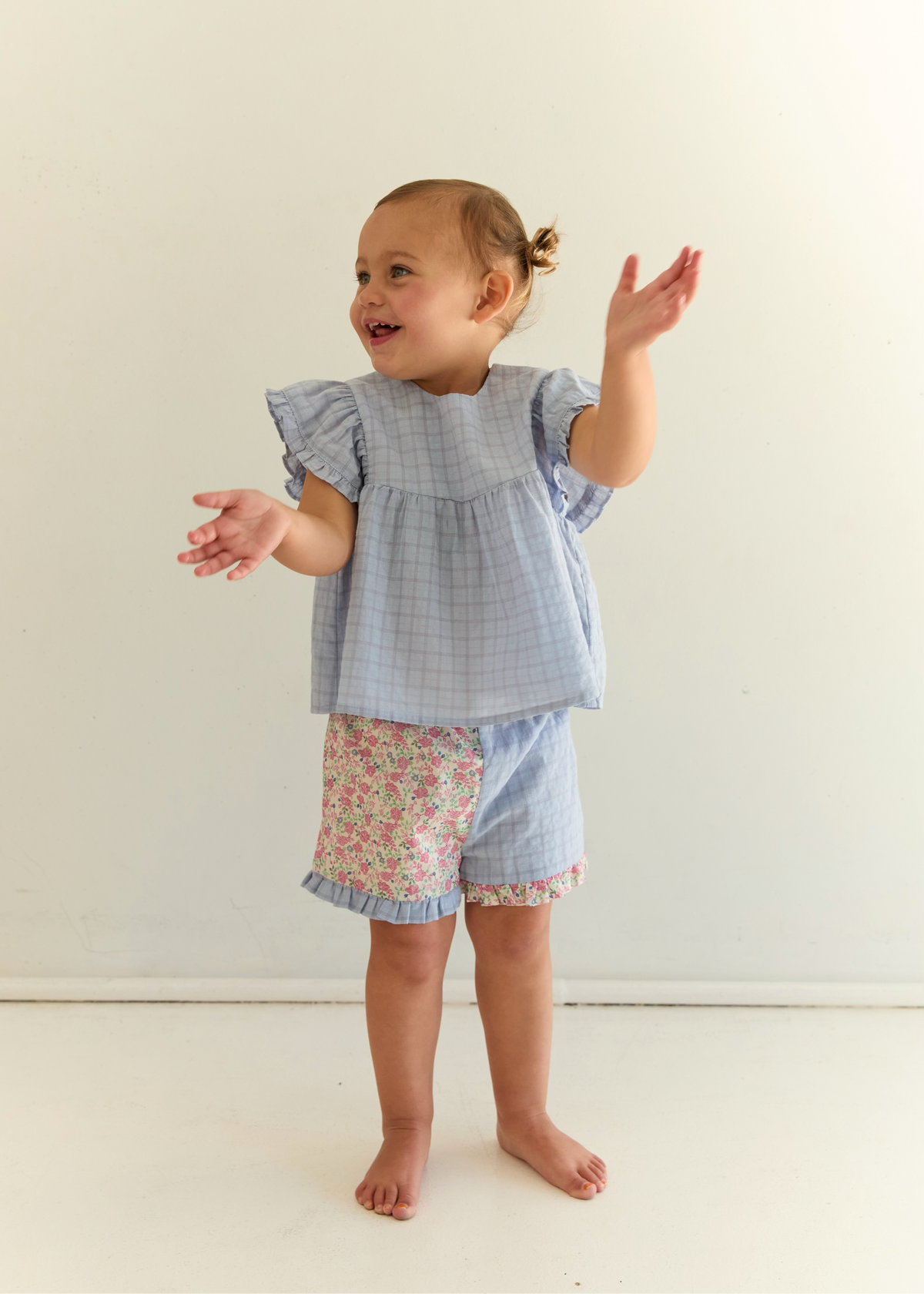 A toddler girl stands barefoot against a plain off-white wall, wearing a light blue and white gingham ruffle-sleeve top and matching shorts with a pink floral patch pocket.