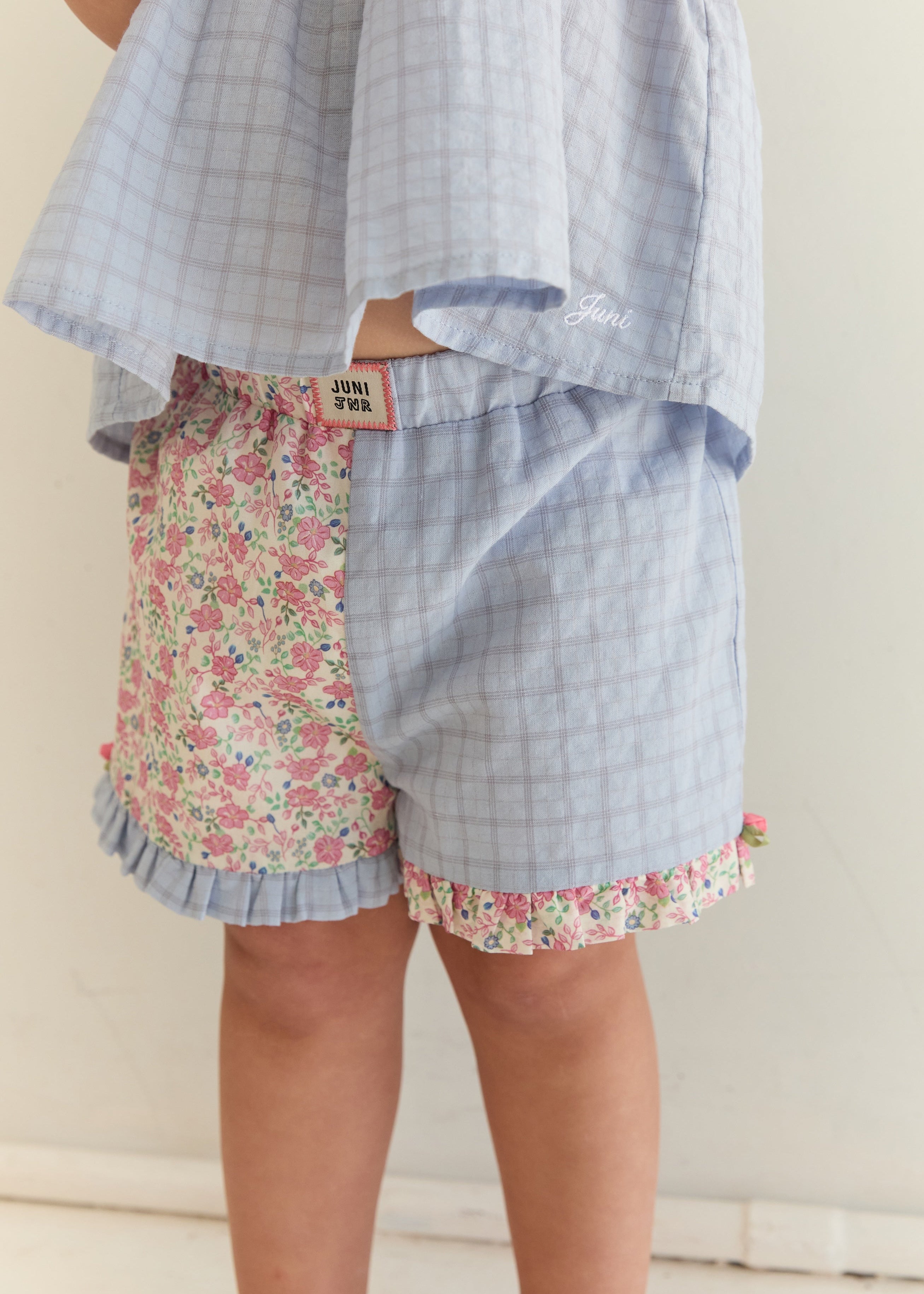 A child wearing a matching two-piece outfit with a light blue gingham top and shorts, paired with floral print shorts on the left leg and ruffled trim.