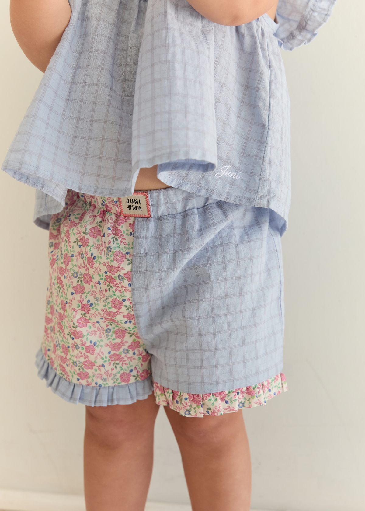 A child wearing a matching two-piece outfit with a light blue gingham top and shorts, paired with floral print shorts on the left leg and ruffled trim.